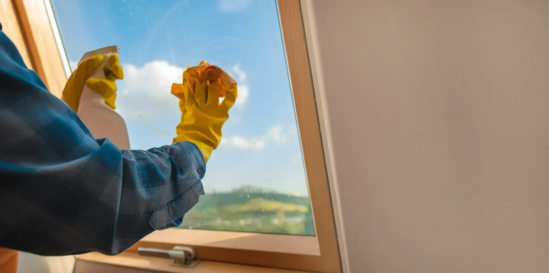 A person wearing yellow gloves sprays and wipes a window, with a view of a blue sky and trees outside.