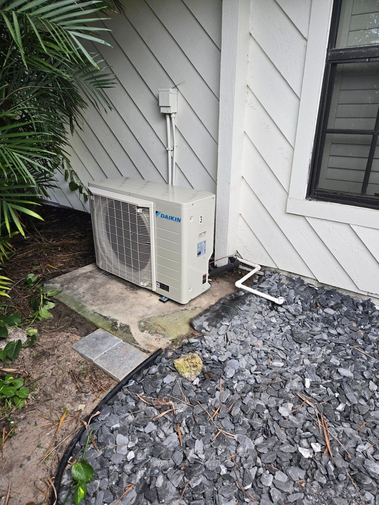 A white air conditioner is sitting on the side of a house next to a window.