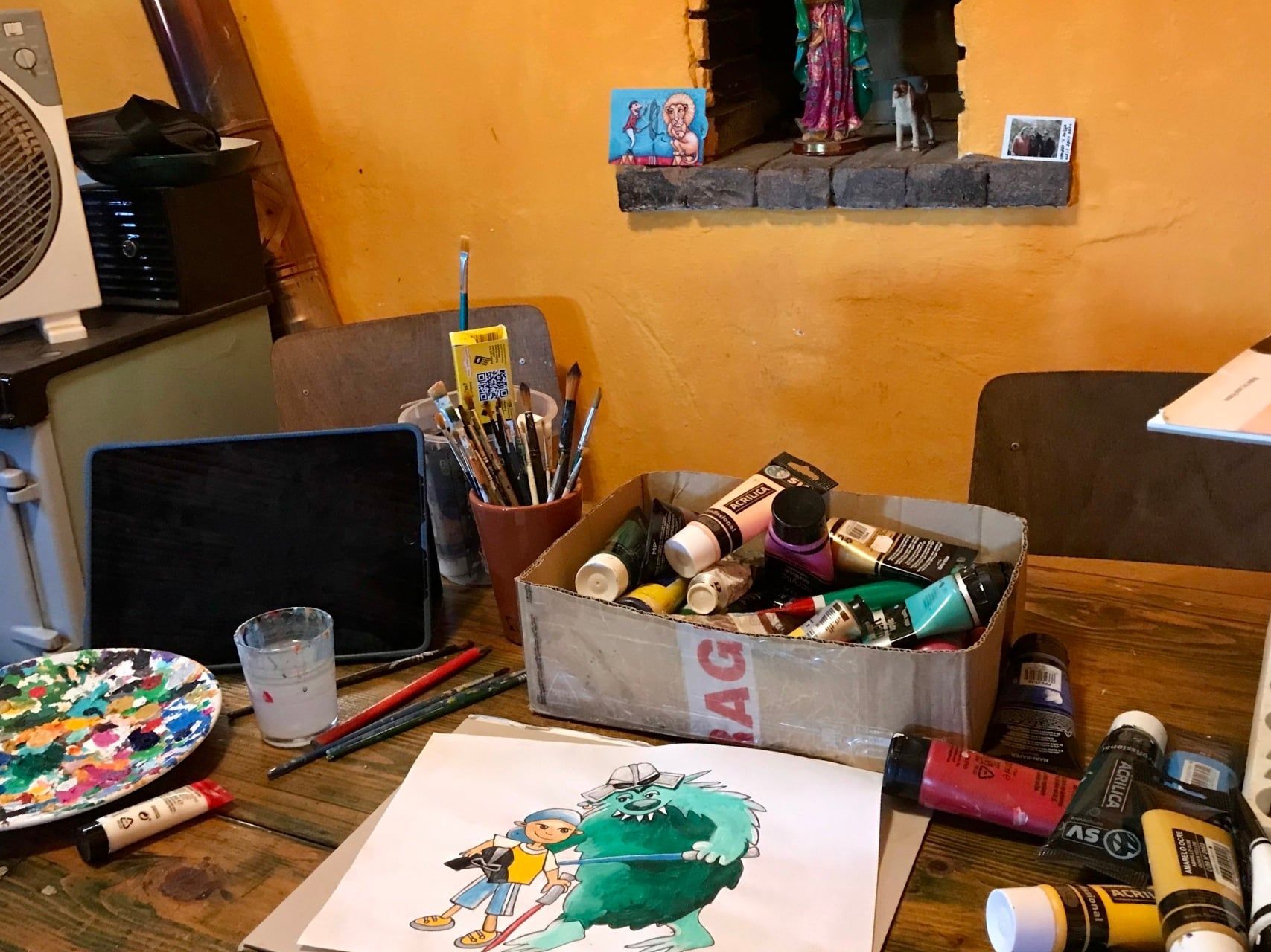 Een kunstenaarswerkplek met schildersbenodigdheden, een laptop, penselen en aquarelverf op een houten bureau.