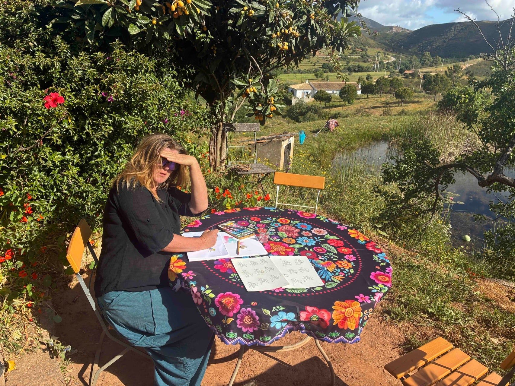Vrouw zit aan een kleurrijke buitentafel en leest de krant in een zonnige tuin aan het water met uitzicht op de bergen.