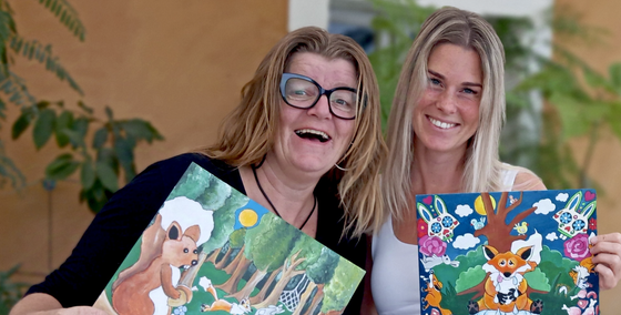Twee lachende vrouwen houden kleurrijke, geïllustreerde boeken vast tegen een groene, binnenhuisomgeving.