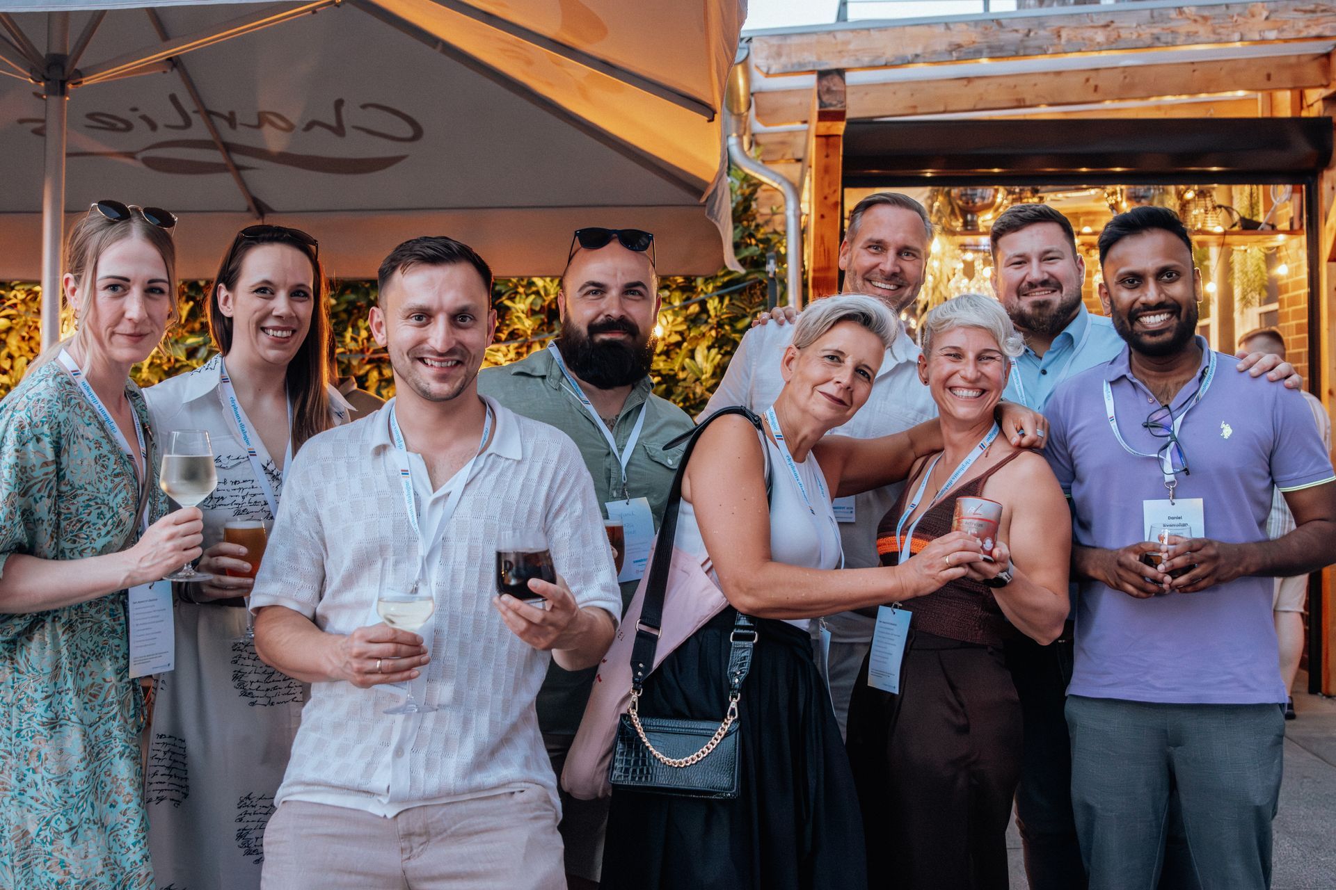 Eine Gruppe von Menschen unterhält sich bei Getränken im Freien auf einer Terrassenparty unter einem Baldachin und Lichterketten.