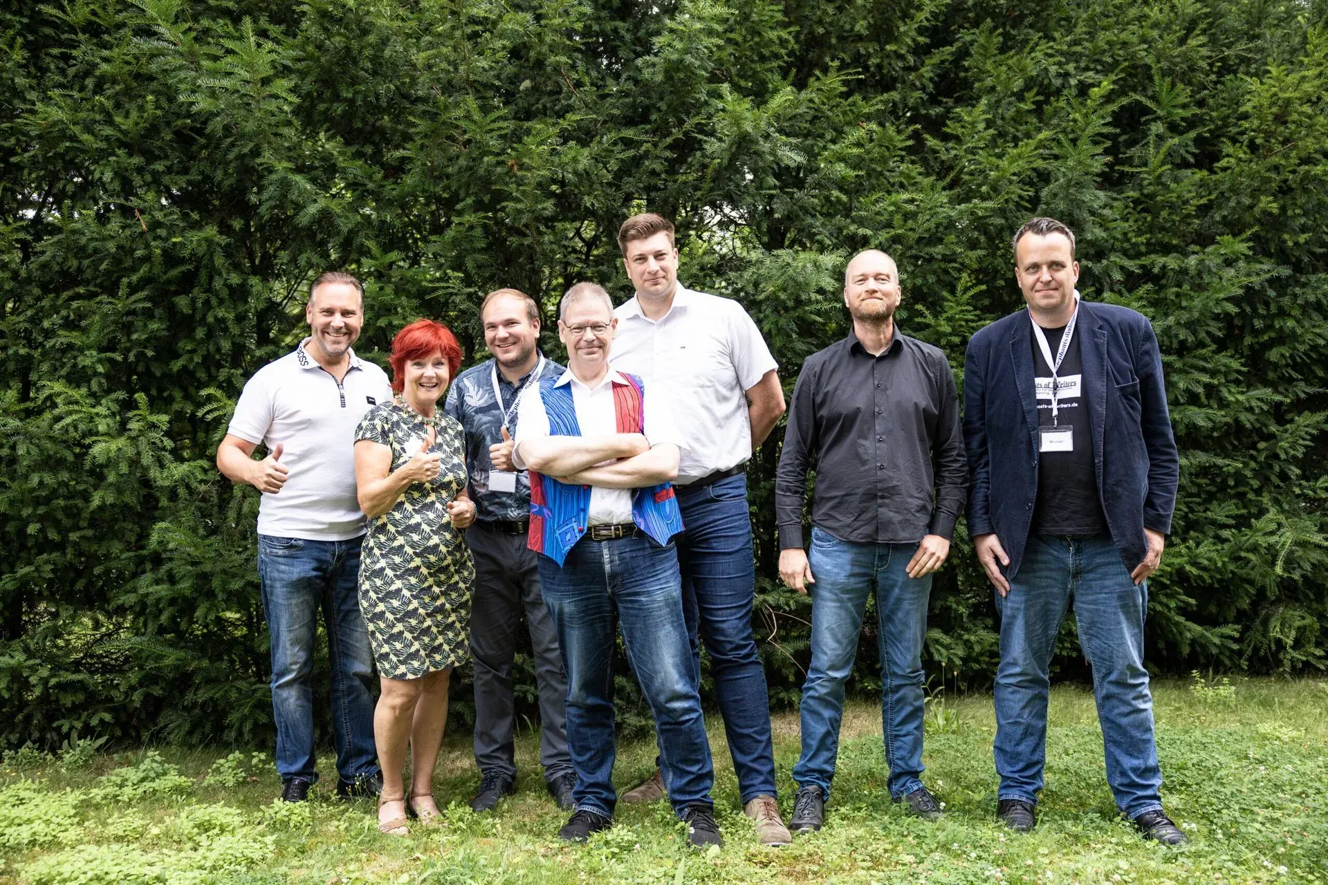 Eine Gruppe von sieben Personen posiert vor grünen Bäumen. Sie lächeln und tragen legere Kleidung, meist Jeans.