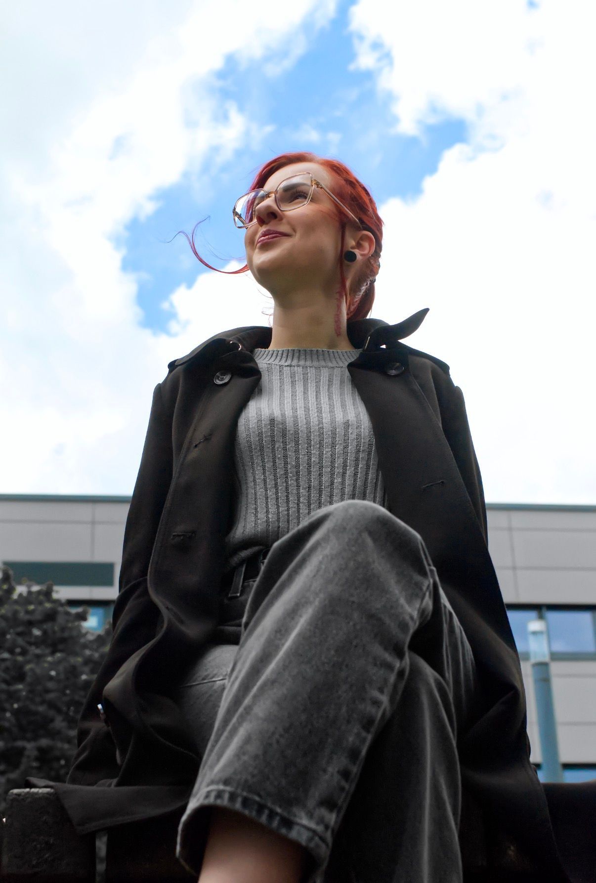 Eine rothaarige Person sitzt im Freien vor einem bewölkten Himmel und trägt eine schwarze Jacke und einen grauen Pullover.