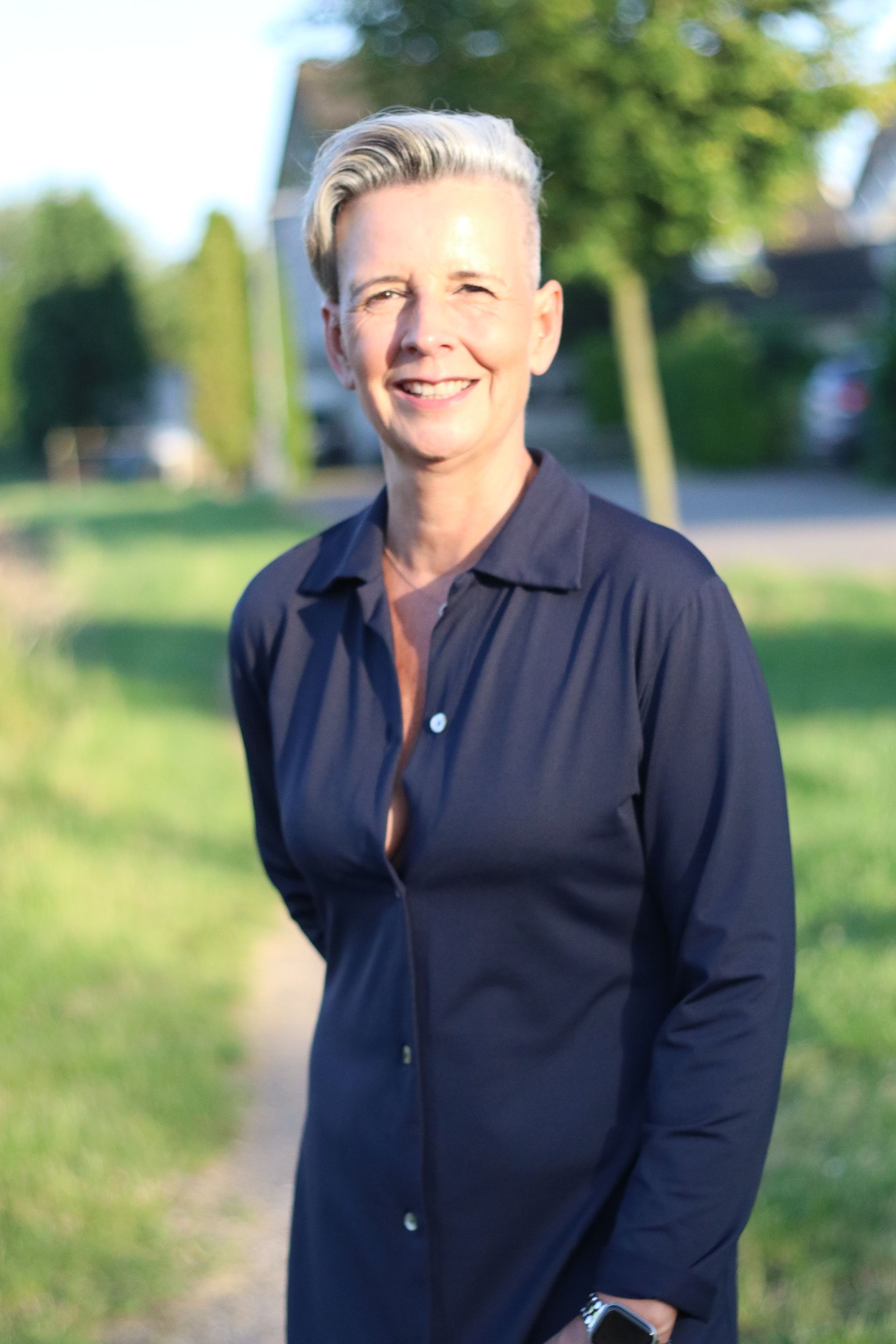 Eine lächelnde Person in einem dunkelblauen Hemd steht im Freien auf einem sonnigen Weg vor dem Hintergrund grüner Bäume.