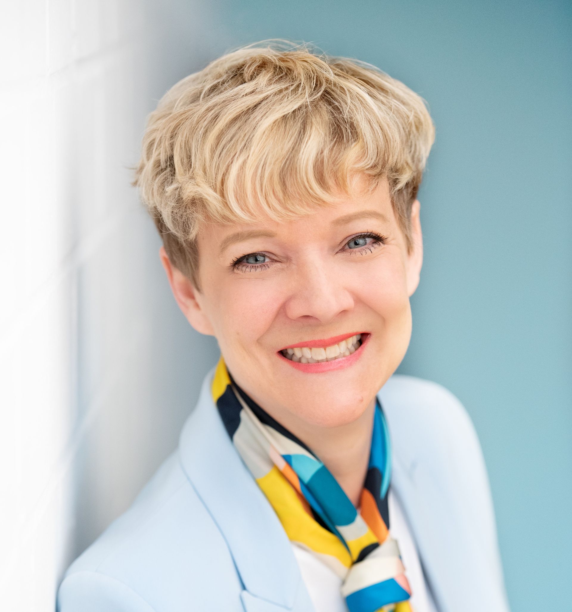 Eine lächelnde Frau in einem hellblauen Blazer und einem bunten Schal lehnt an einer Wand.