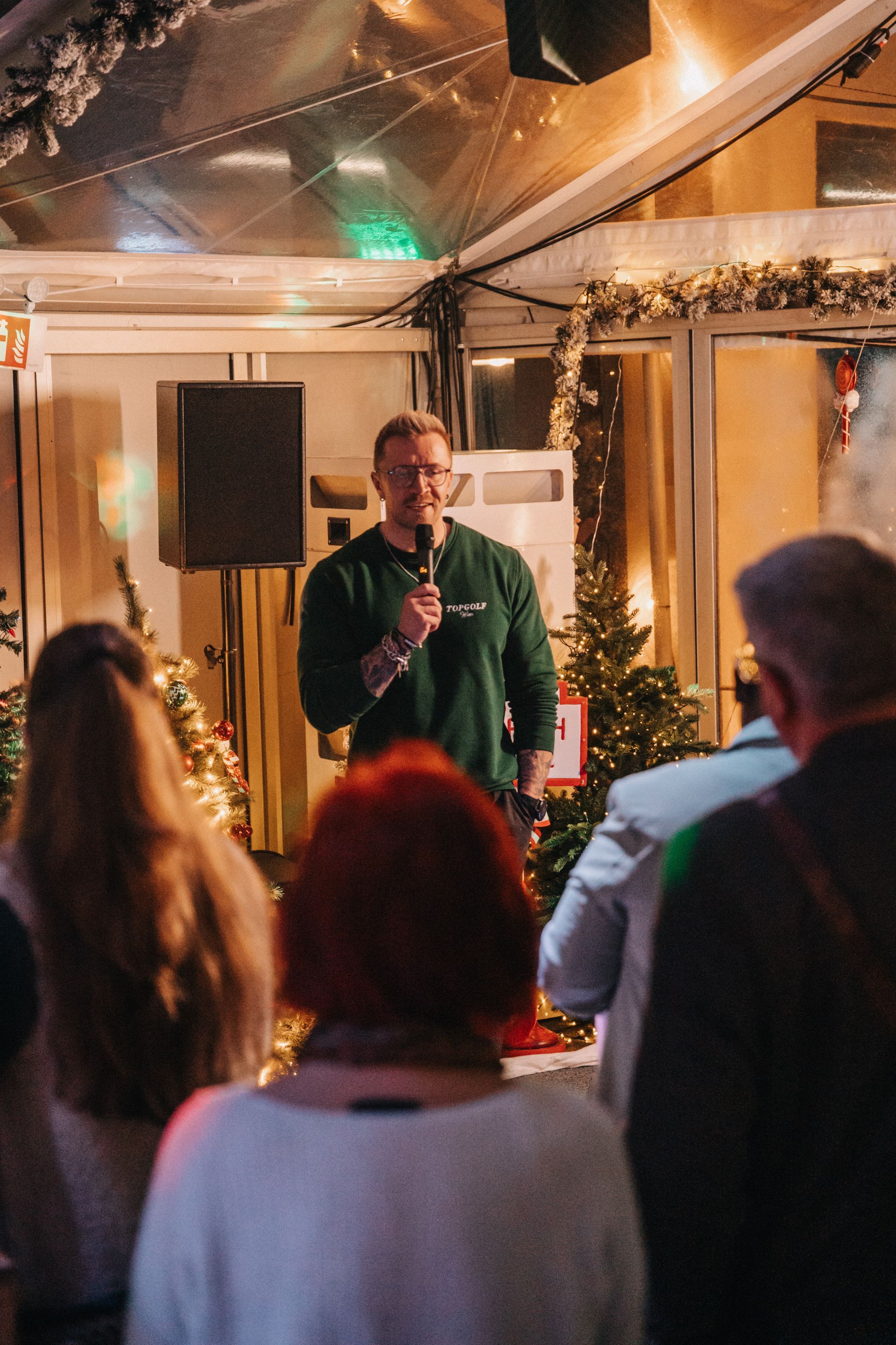 Ein Mann spricht bei einer Veranstaltung in ein Mikrofon, umgeben von Menschen und Weihnachtsdekorationen.