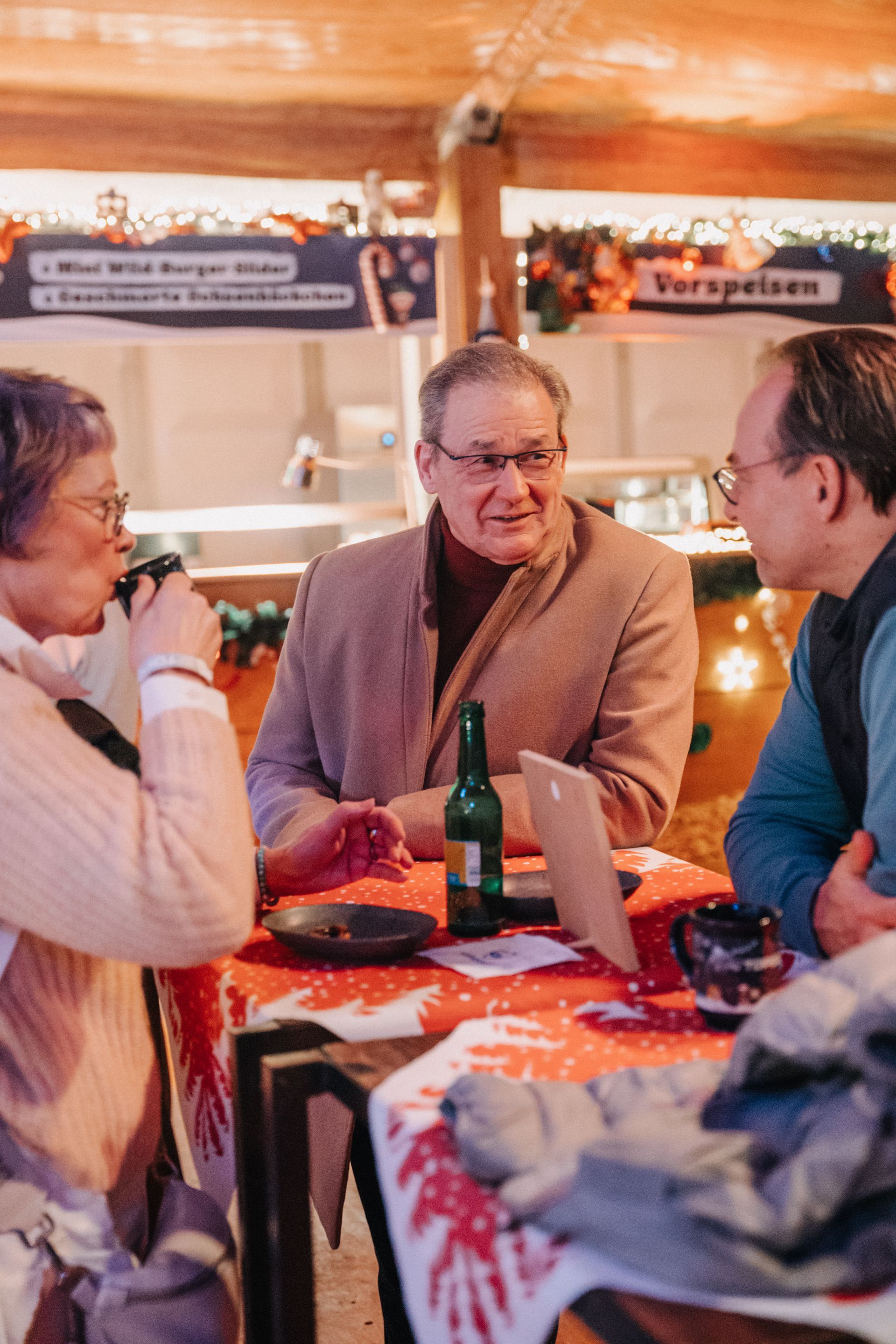 Drei Personen sitzen an einem Tisch, trinken und unterhalten sich, Weihnachtsmarktkulisse, rote Tischdecke.