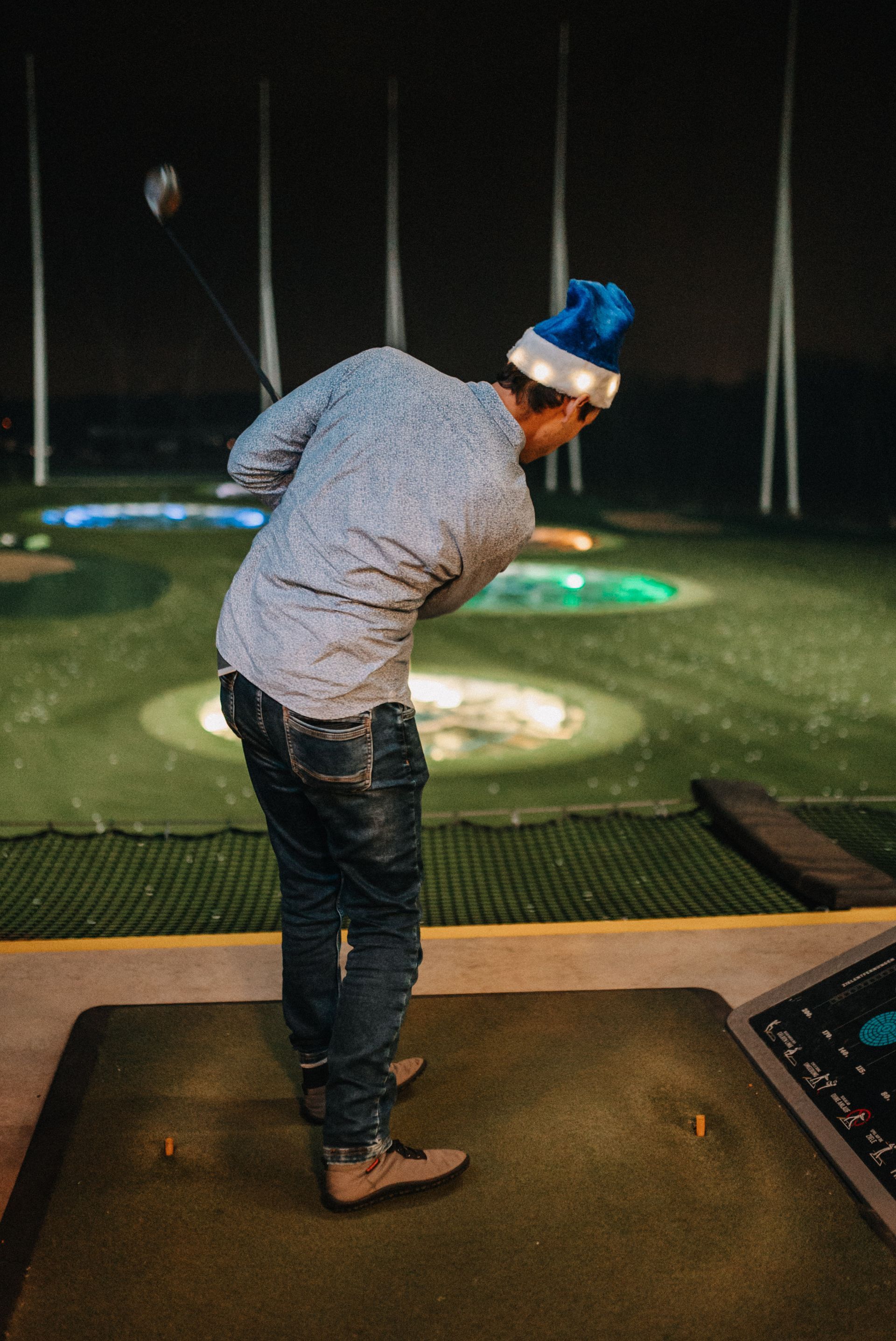 Eine Person, die auf einer nächtlichen Driving Range mit einem Golfschläger schwingt und einen blauen Hut mit Lichtern trägt.