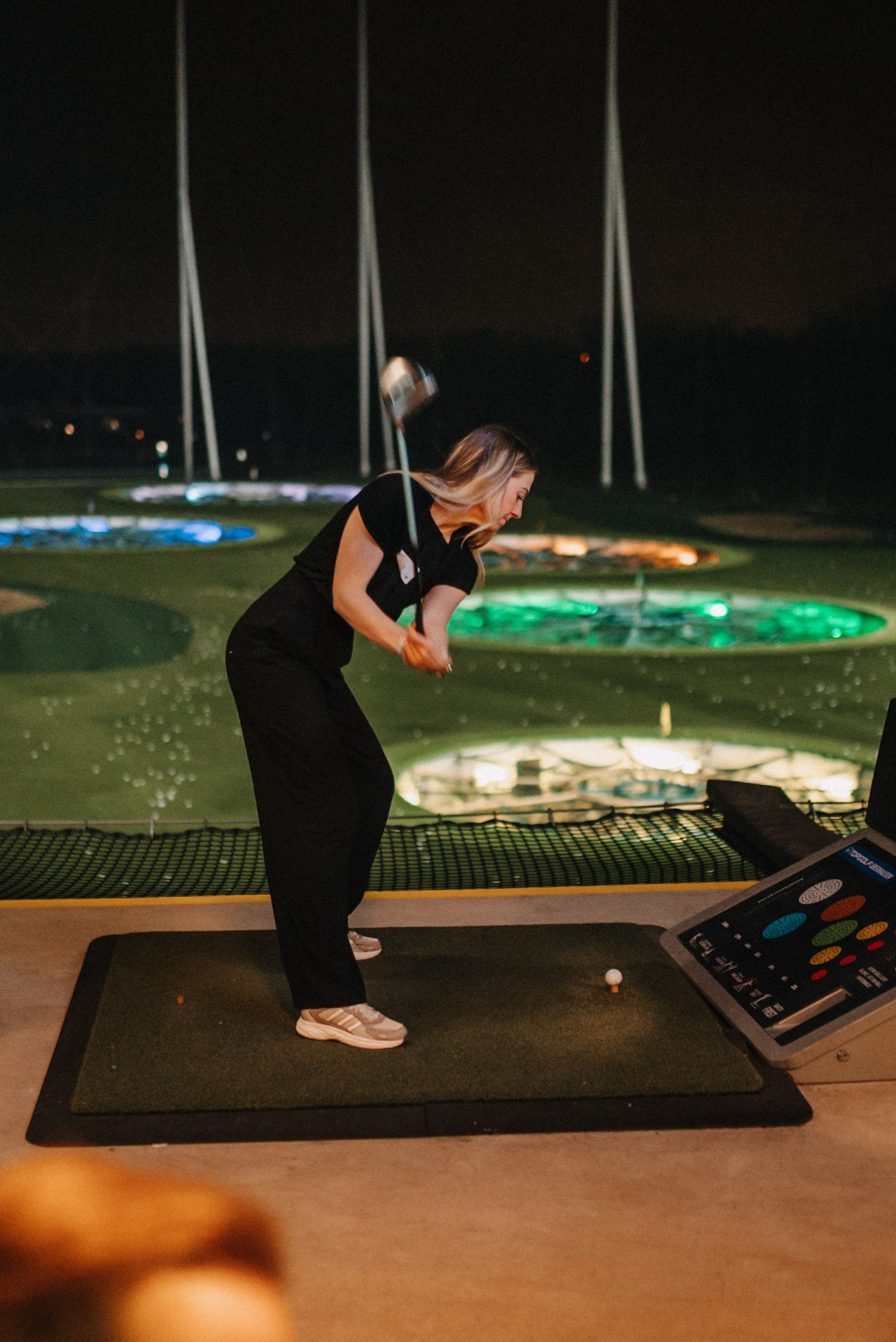 Eine Frau schwingt einen Golfschläger auf einer nächtlichen Driving Range mit leuchtenden Zielscheiben.