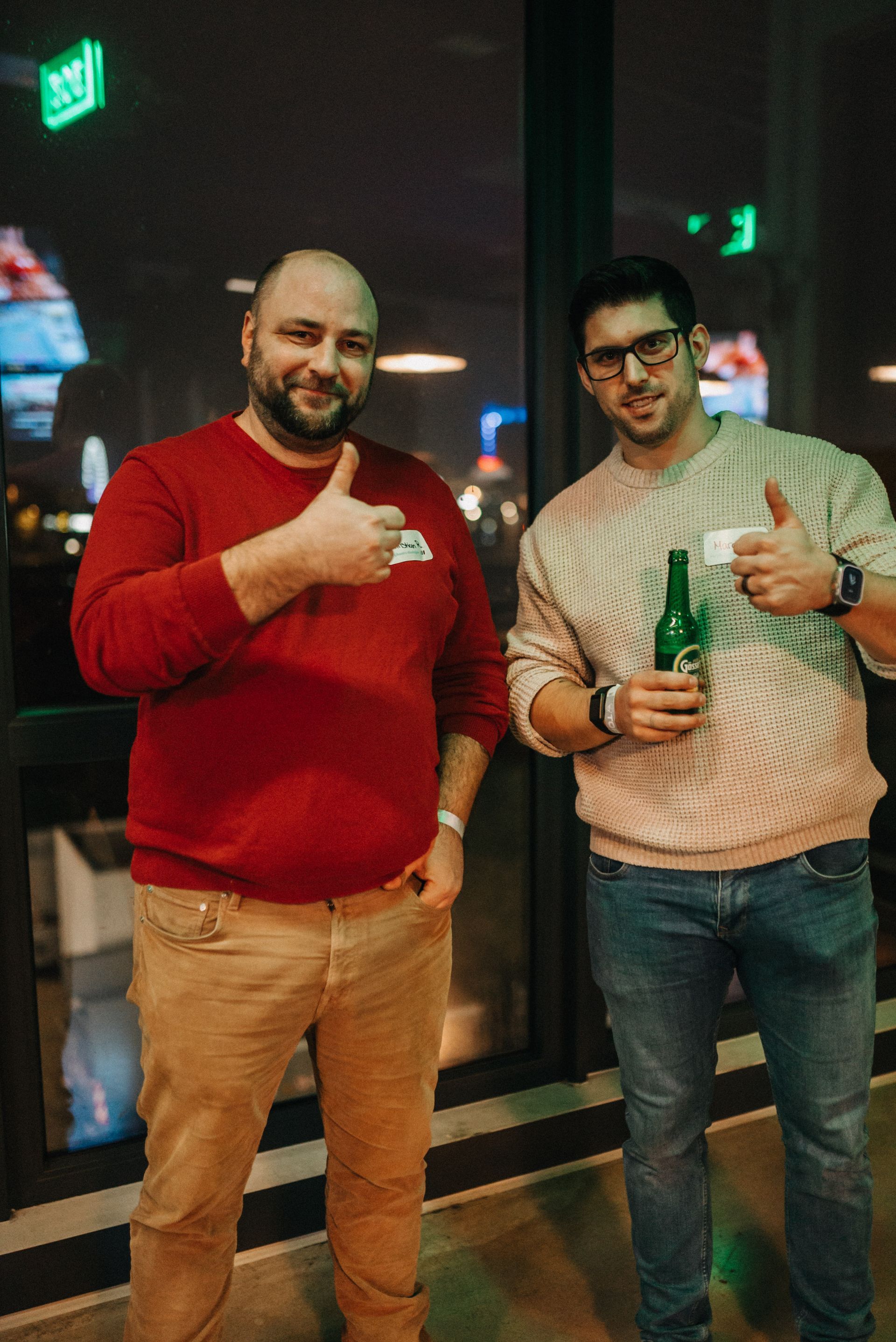 Zwei Männer, einer in einem roten, der andere in einem cremefarbenen Pullover, zeigen den Daumen nach oben. 