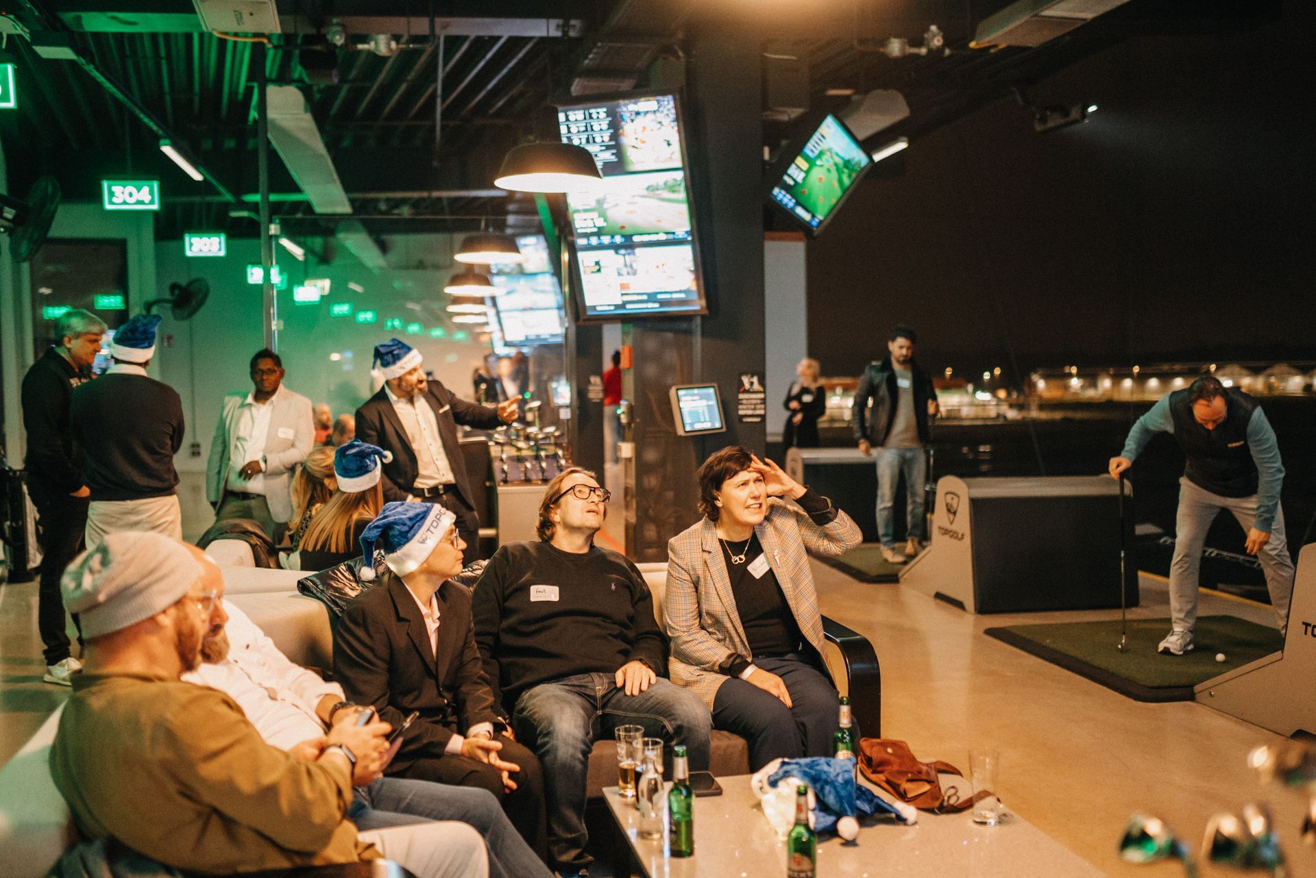 In einer Indoor-Golfanlage schauen sich Leute auf Bildschirmen Golf an. Einige tragen Weihnachtsmannmützen.
