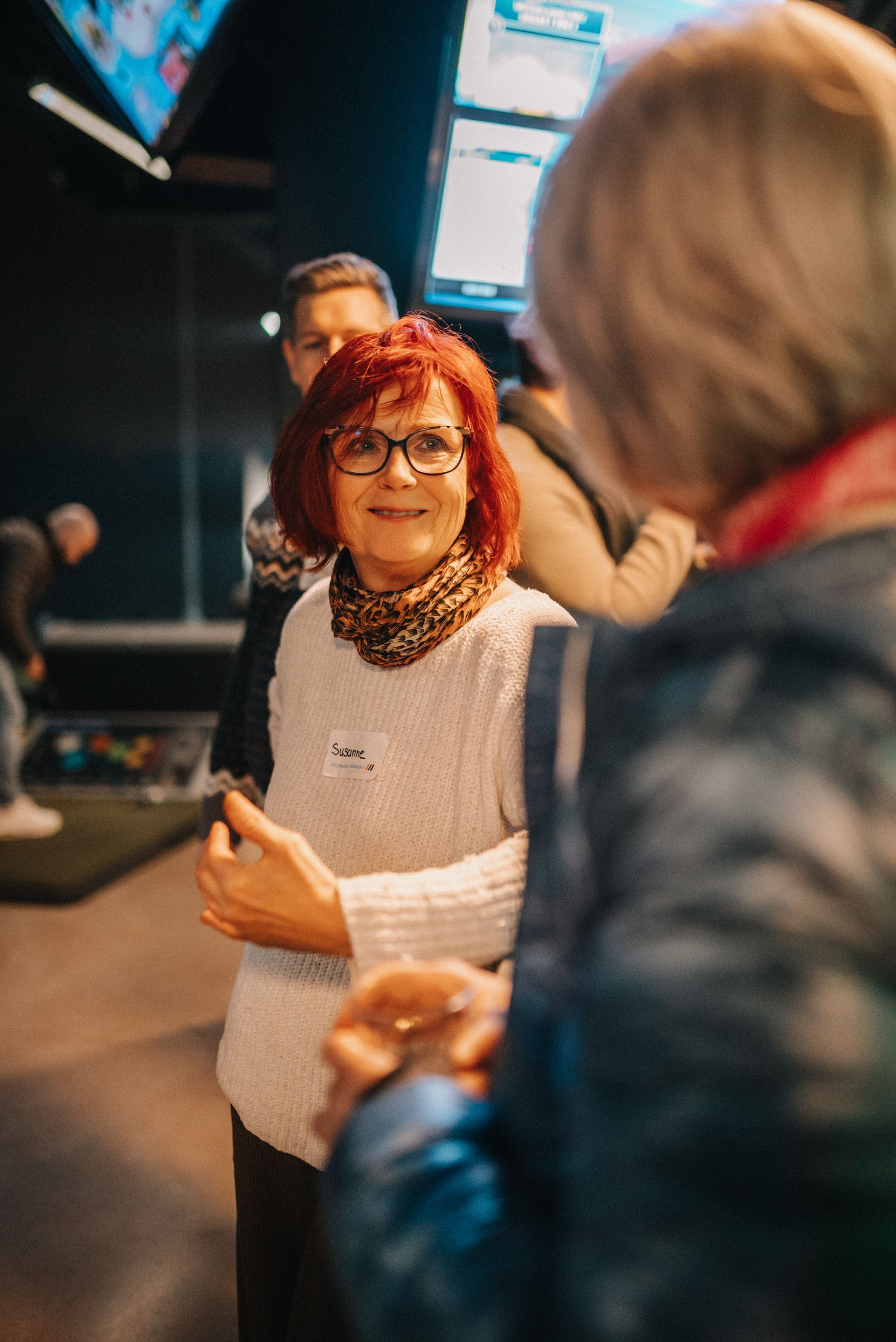 Eine rothaarige Frau mit Brille lächelt während eines Gesprächs bei einer geselligen Zusammenkunft.