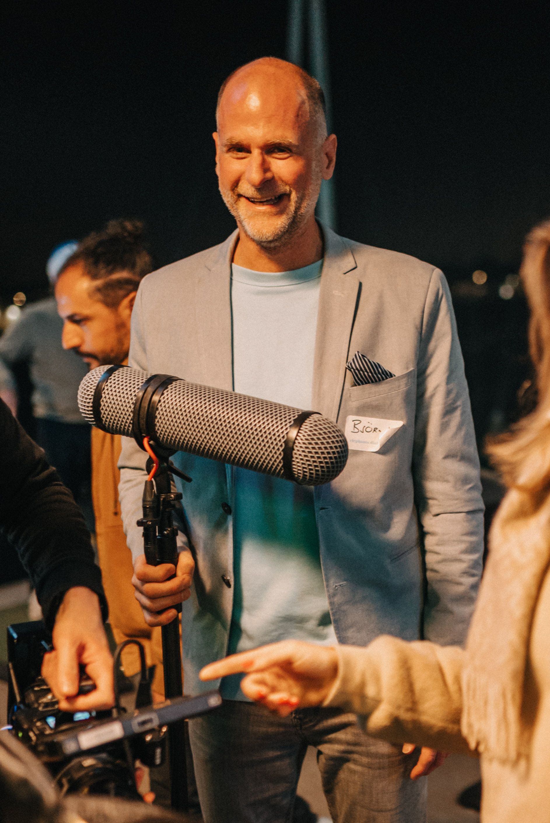 Ein Mann mit Mikrofon in der Hand lächelt. Er trägt einen hellen Blazer und ein blaues Hemd und steht nachts im Freien.