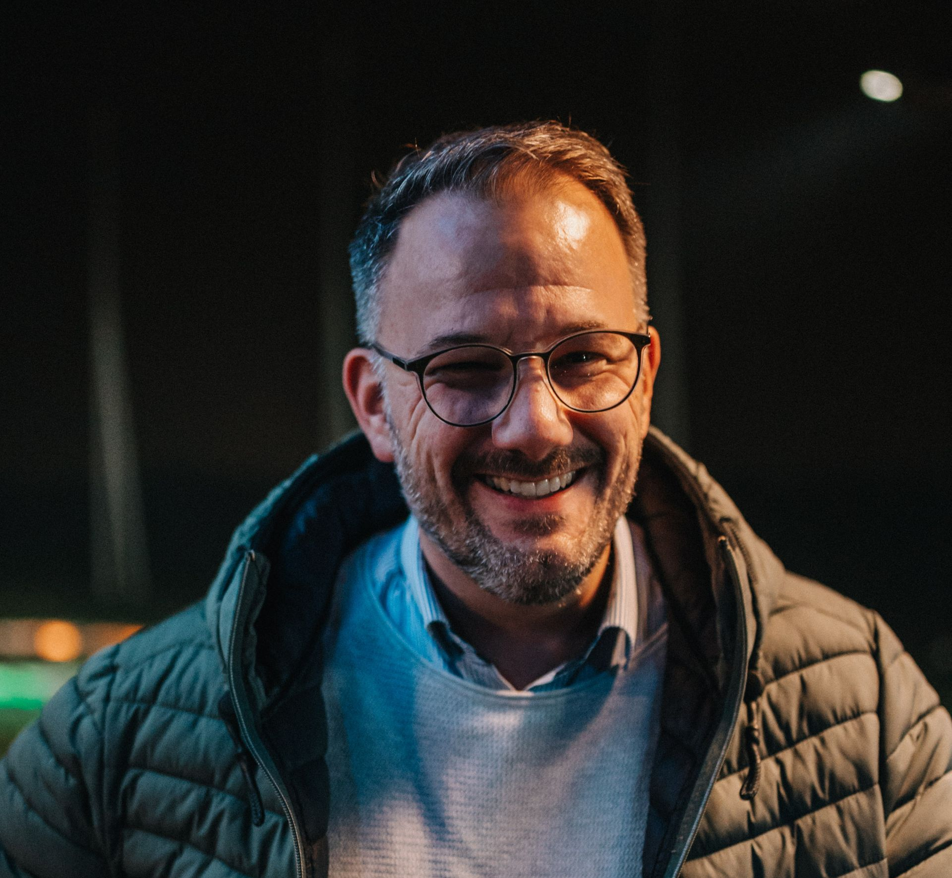 Ein Mann mit Brille lächelt. Er trägt eine grüne Steppjacke und einen hellgrauen Pullover. Dunkler Hintergrund.