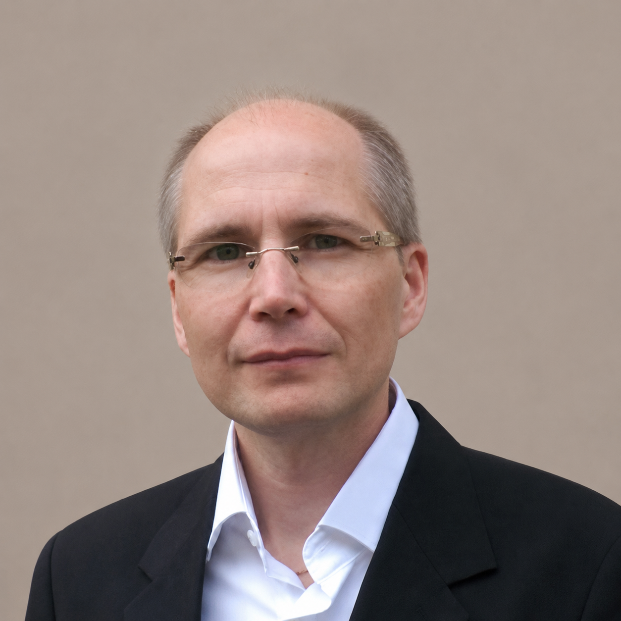 Porträt eines Mannes mit Brille, der einen dunklen Anzug und ein weißes Hemd trägt, vor einem schlichten beigen Hintergrund