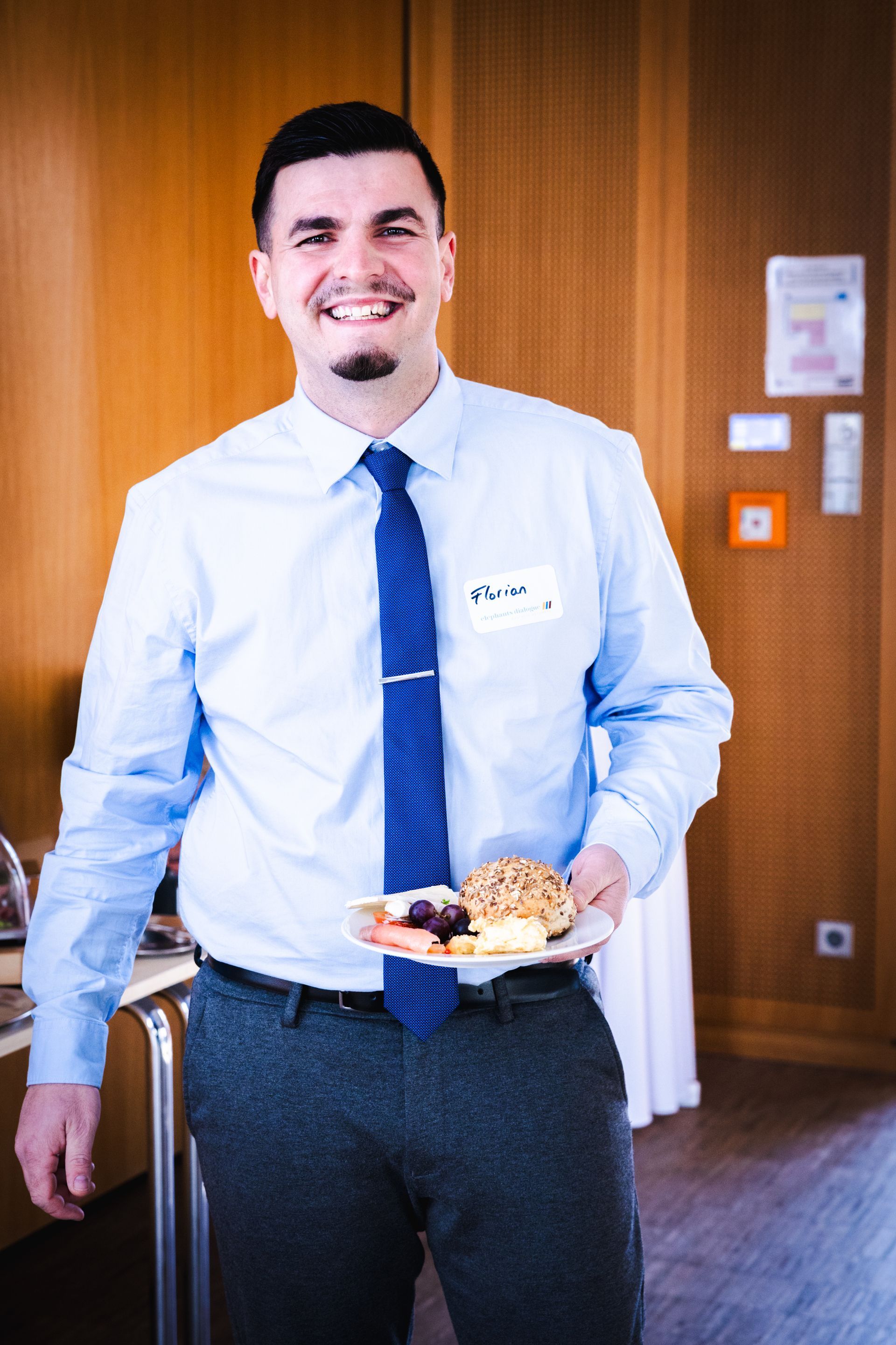 Ein Mann in weißem Hemd und blauer Krawatte hält einen Teller mit Essen in einem holzgetäfelten Raum.