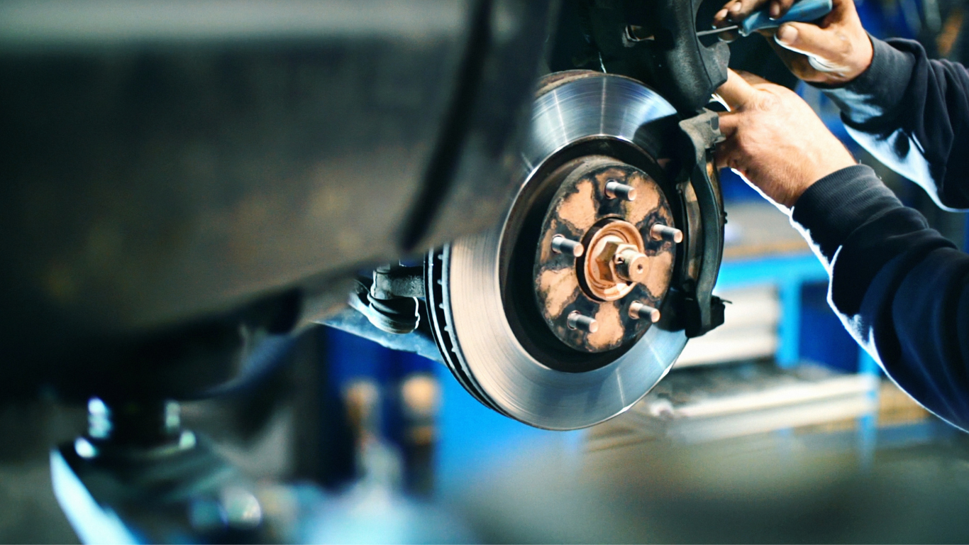 Mechanic in blue overalls examines a white SUV lifted on a hoist in a repair shop.
