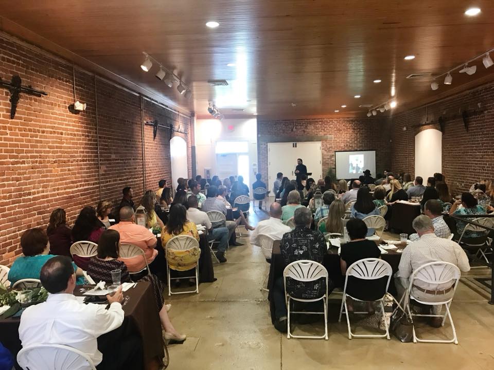Large gathering in a brick-walled room with people seated at tables, listening to a speaker near an open doorway.
