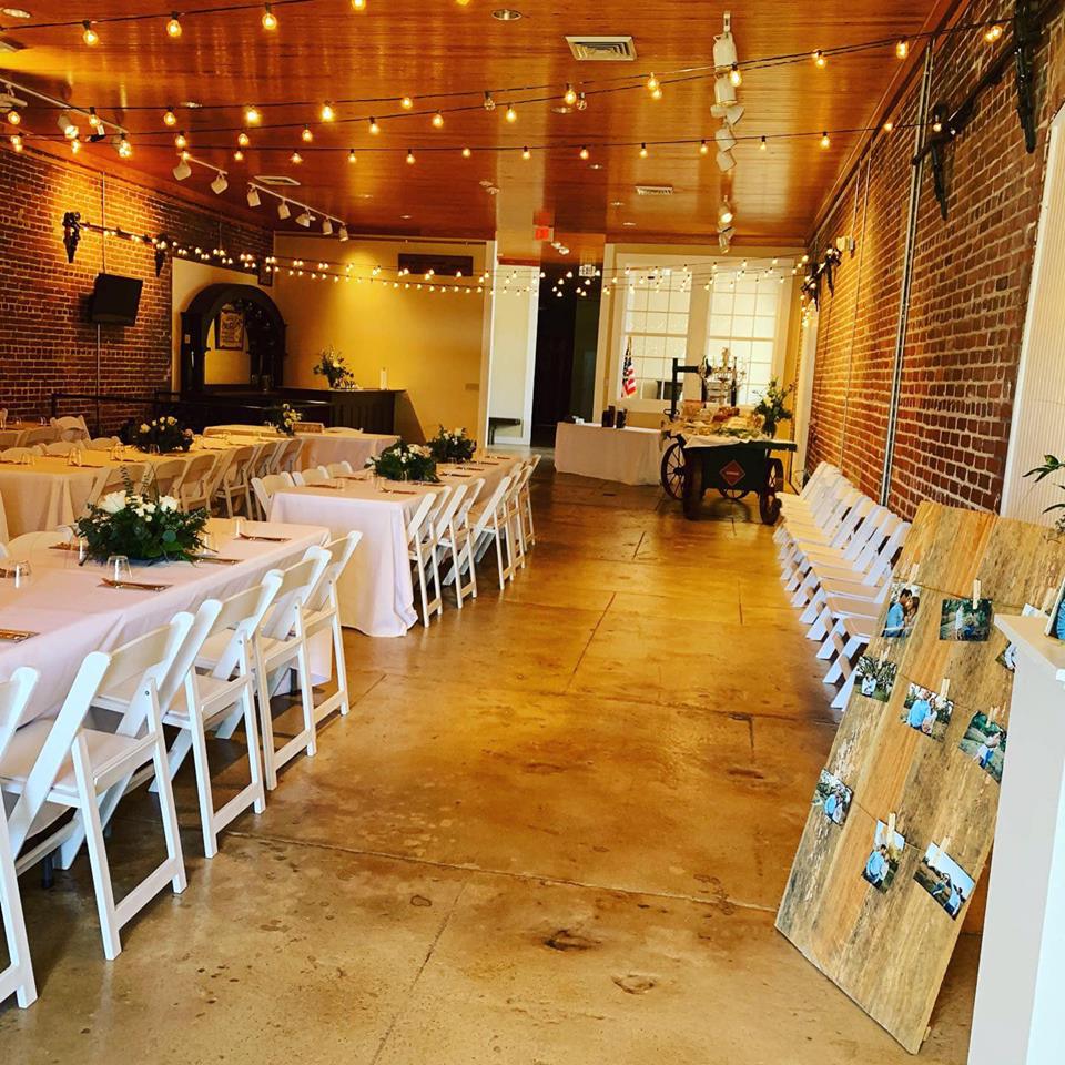 Wedding reception venue with long tables, white chairs, string lights, and exposed brick walls.