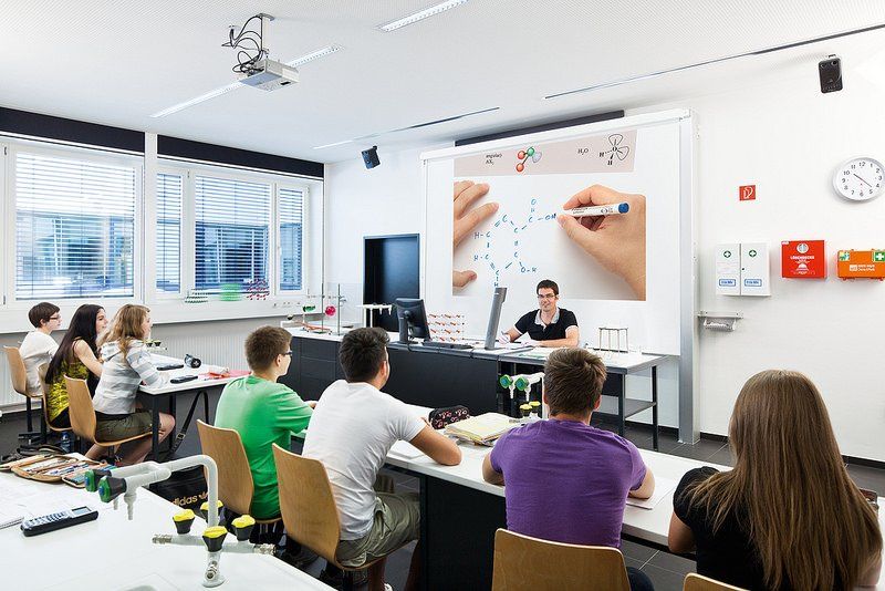 Students watching a teacher demonstrate on a screen in a classroom. The teacher is writing on the screen with blue marker.