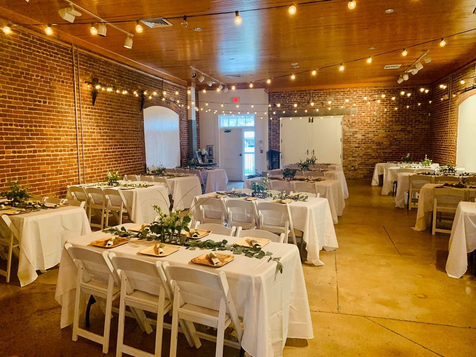 Reception hall set for an event, featuring tables with white linens, string lights, and exposed brick walls.