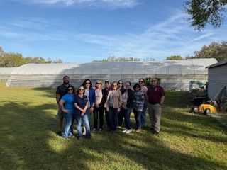 Group of people posing in a grassy field, in front of a large greenhouse structure, under a clear blue sky.