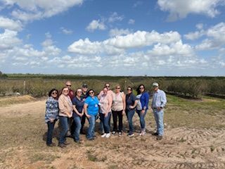 Group of people standing in a field in front of rows of trees, under a cloudy blue sky.