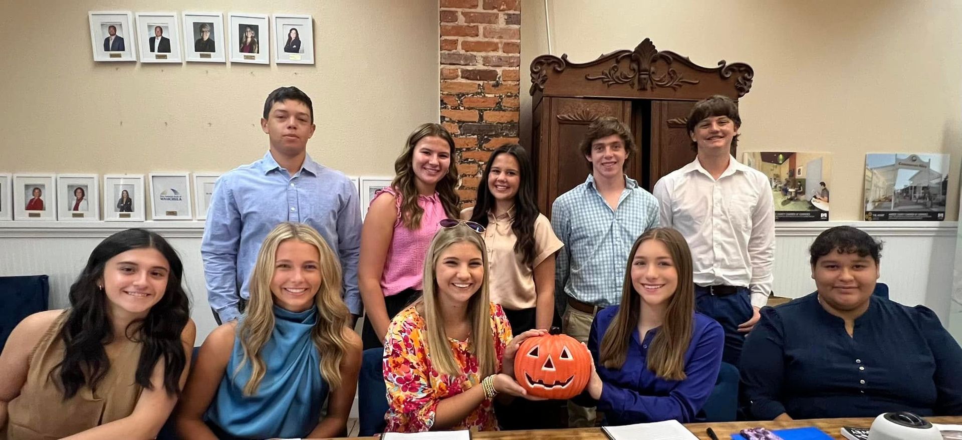 A group of young people smiles at the camera, some holding a small jack-o'-lantern, with a brick wall background.