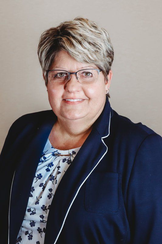 Woman with short, blonde hair and glasses smiles, wearing a blue blazer over a patterned blouse.