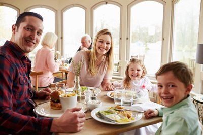 Family smiling at the camera while seated at a restaurant table. Dad wears a plaid shirt; mom and children are beside him. Windows are in the background.