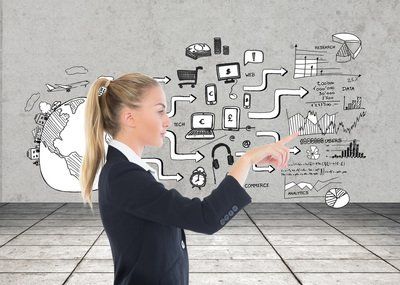 Blonde businesswoman pointing at a wall covered in black-and-white business-related diagrams and doodles in a gray room.