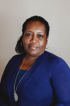 A Black woman with dark hair in a low ponytail, wearing a blue cardigan and a pendant necklace, smiles at the camera.
