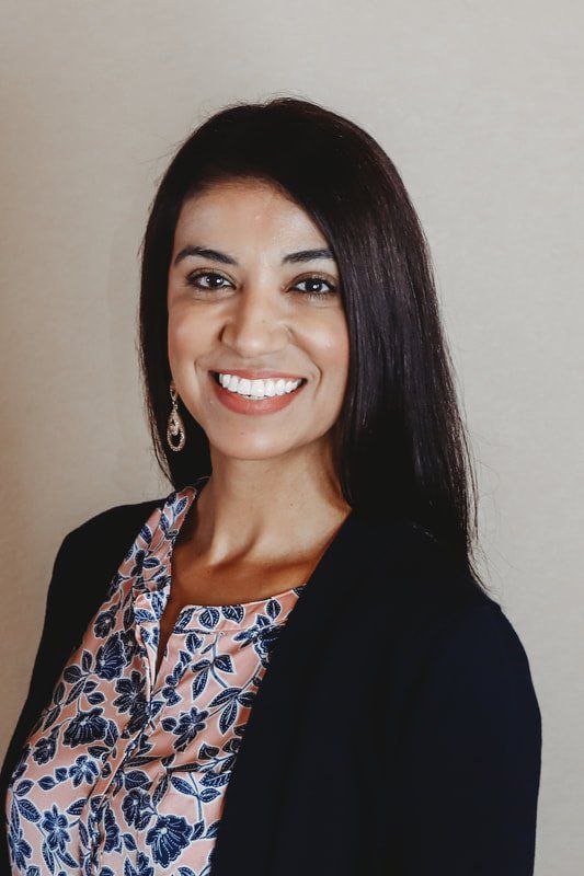 Woman with dark hair smiles. She wears a pink floral top under a black blazer; background is beige.