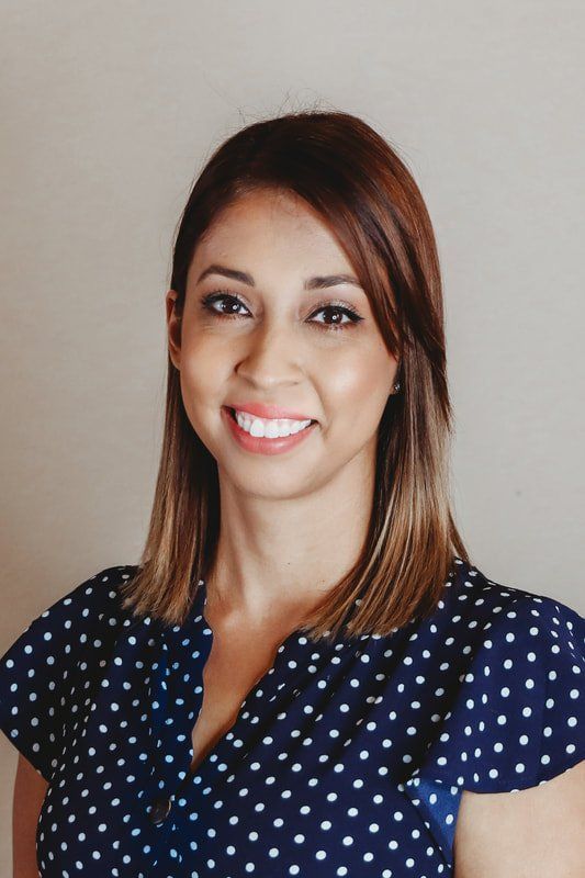 Woman with brown hair and a polka dot shirt smiles at the camera. She is in front of a neutral wall.