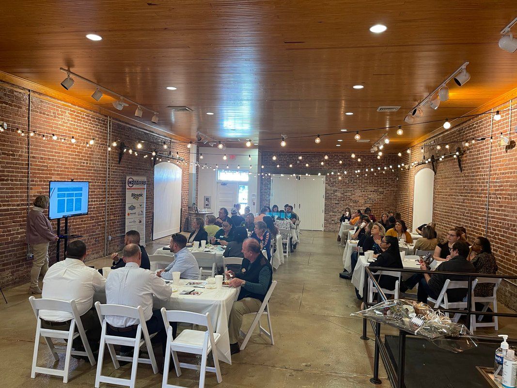 A conference taking place in a brick-walled room. People sit at tables facing a presentation screen. Strings of lights hang from the ceiling.