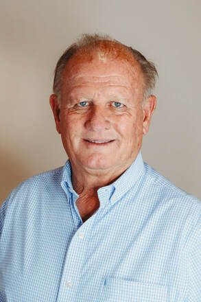 Older man with fair skin, blue eyes, and thinning hair, wearing a light blue button-down shirt, smiling slightly against a neutral background.