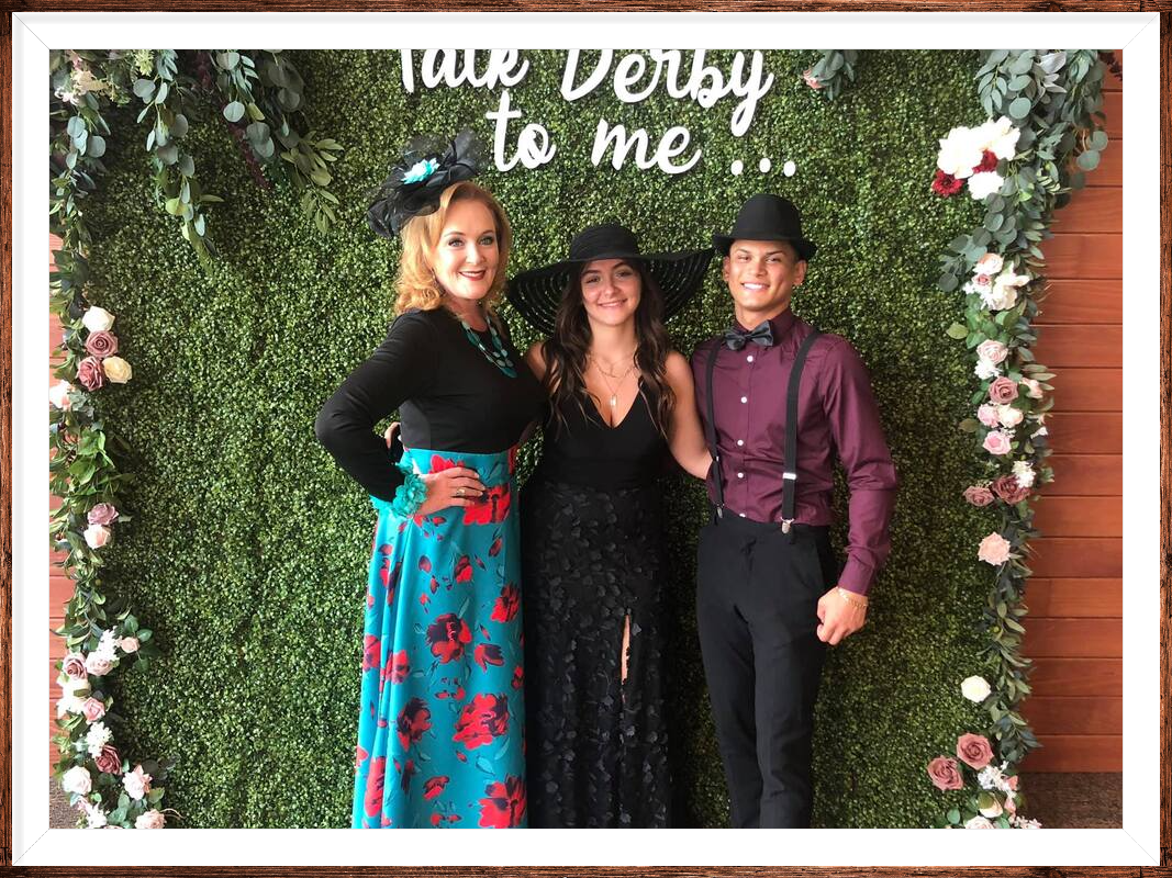 Three people pose in front of a green wall with floral decor. Two women and a man are dressed up, likely at a Derby event.