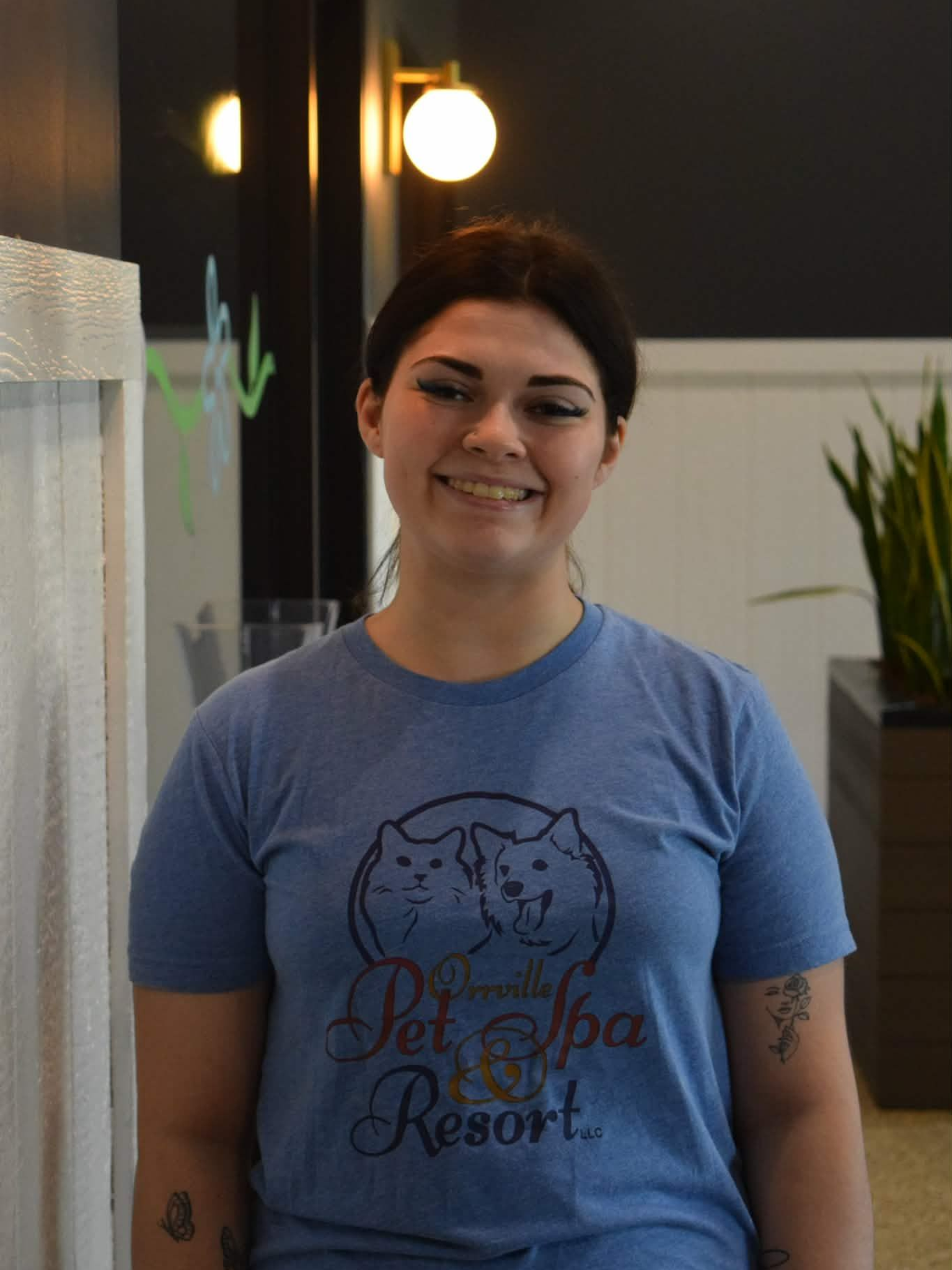 A young woman in a blue sweatshirt smiles at the camera, sitting in a modern hallway with plants and dark ceiling.