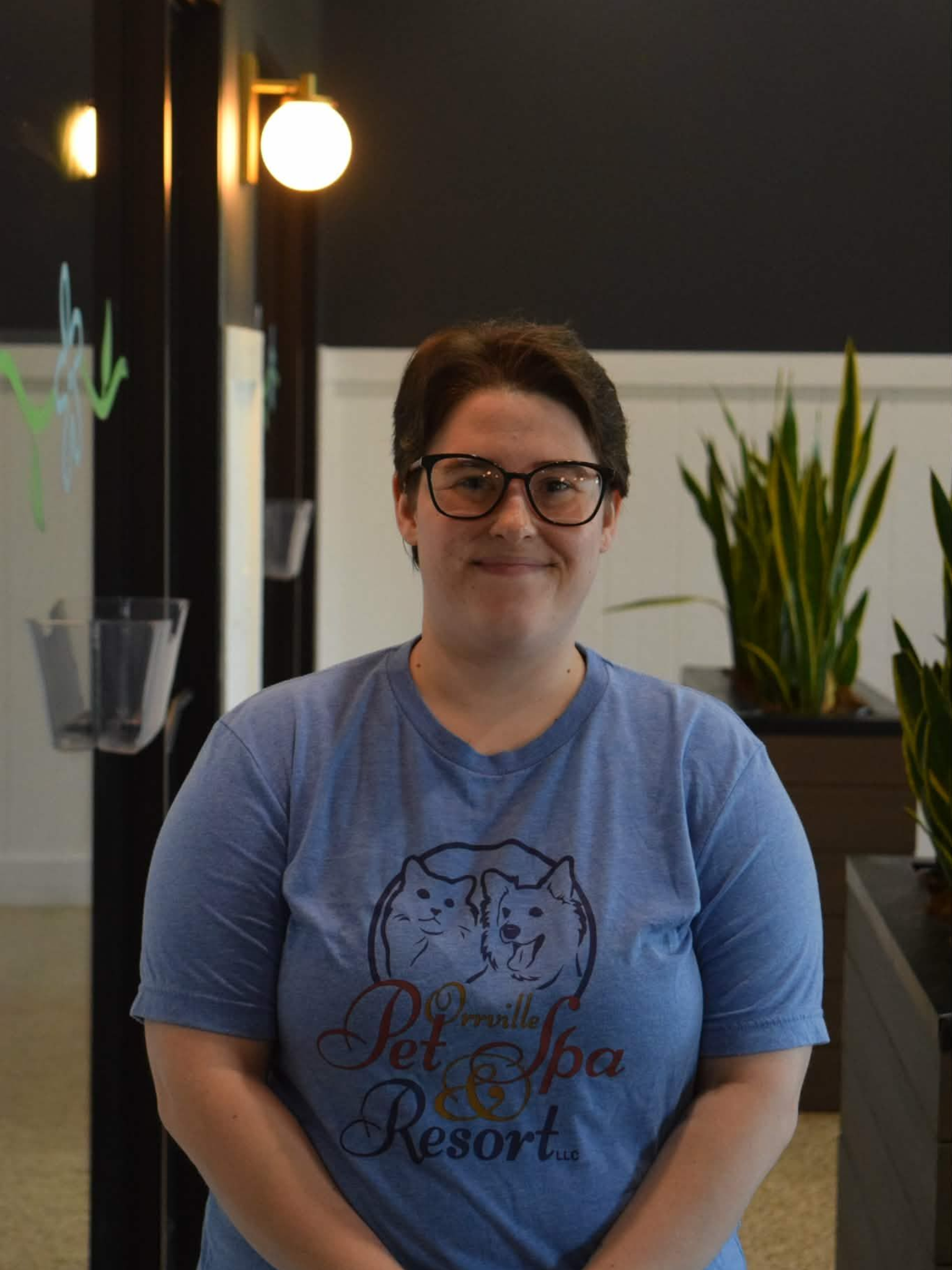 Woman with glasses smiling in a hallway, wearing a blue sweatshirt. Snake plants in wooden planters line the wall behind her.