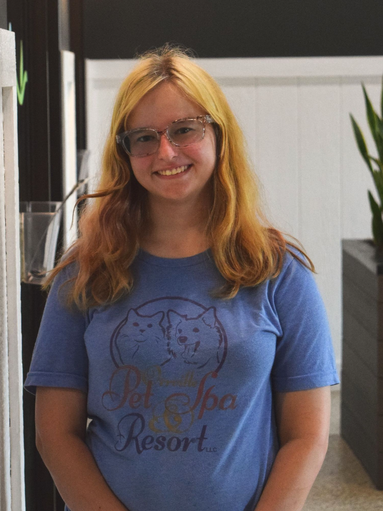 Woman with glasses sits in a wooden chair, wearing a blue shirt with a wolf design and black leggings, indoors. She is smiling.