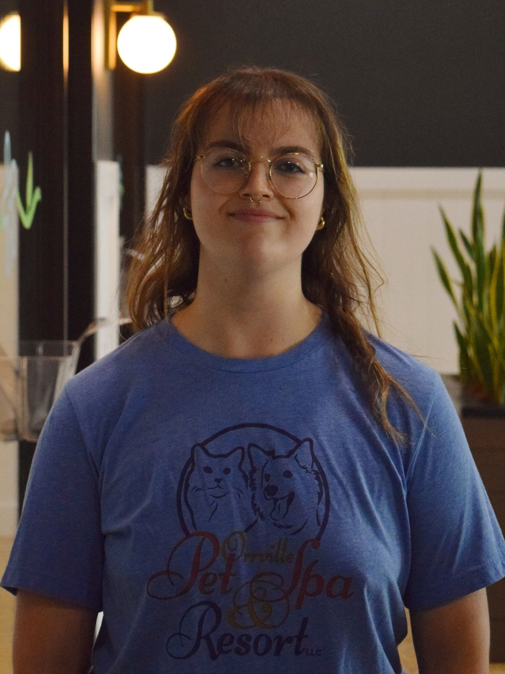 A young woman with red hair and glasses smiles at the camera, wearing a blue sweatshirt. She sits indoors near plants.