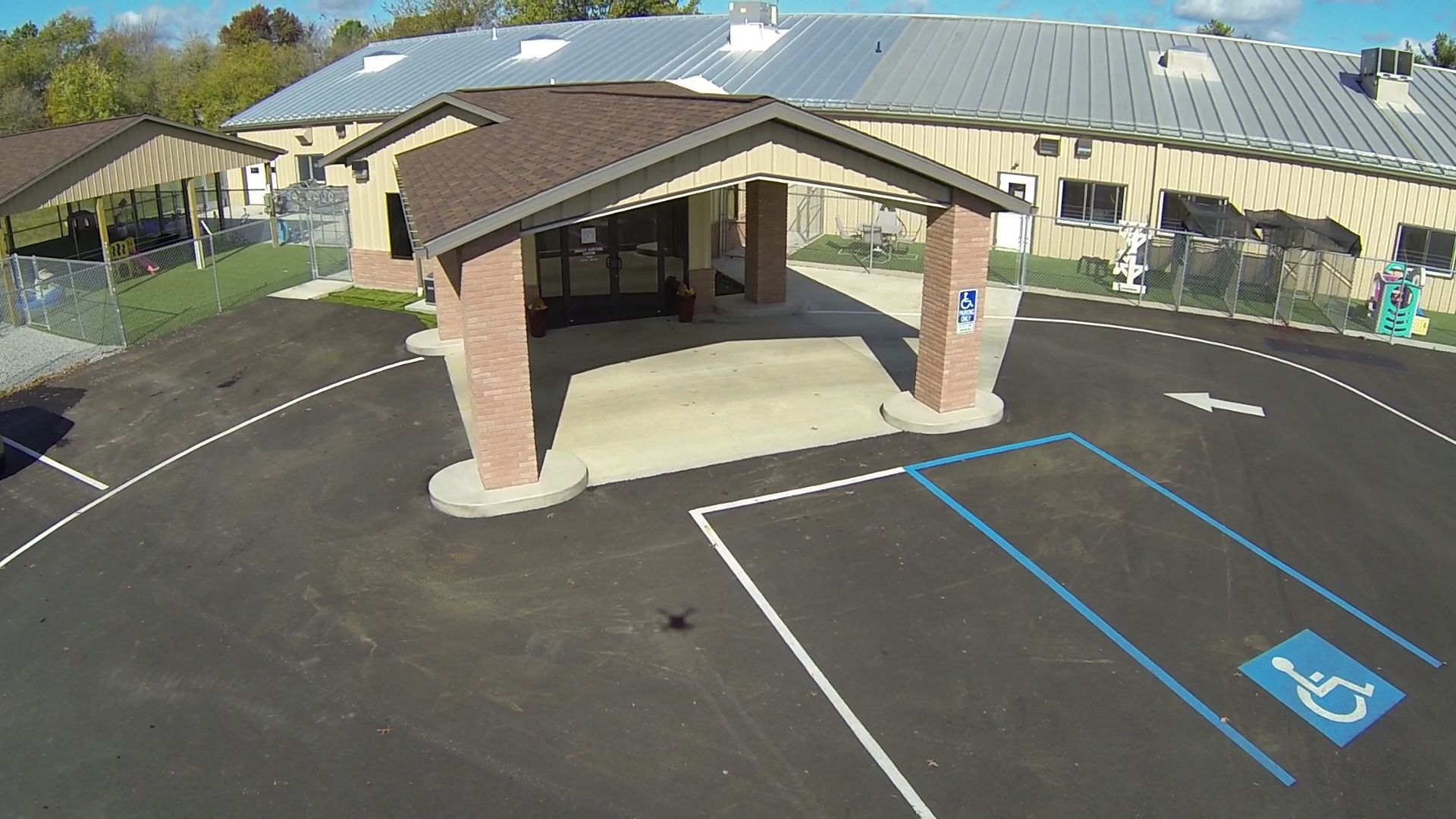 Aerial view of a building with a covered entrance and a handicap parking space. Brown and grey roofing; asphalt driveway.