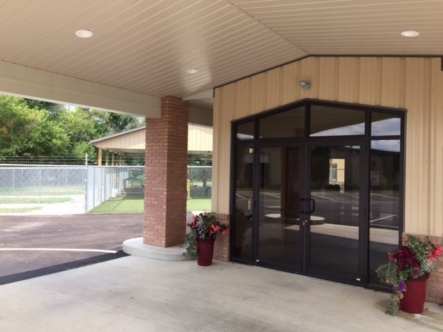 Exterior view of a building with a covered entrance, glass doors, brick pillar, and potted flowers. A fence and other structures are visible in the background.