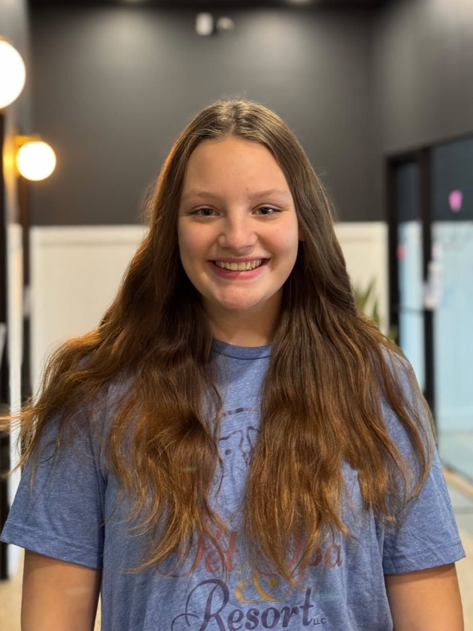Young woman with long blonde hair sitting in a brown chair, wearing a blue t-shirt, smiling. She is in a modern interior with plants.