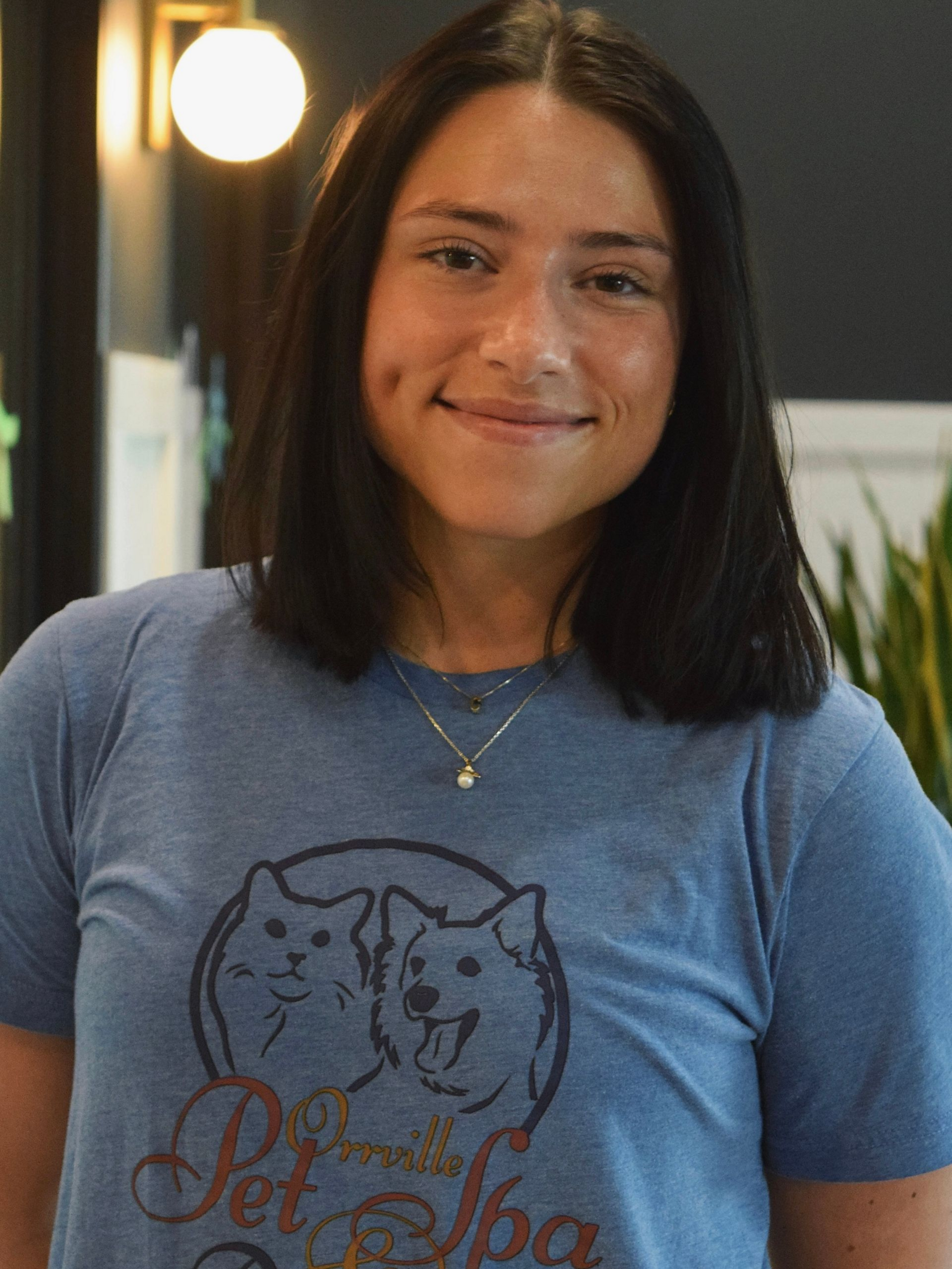 A young woman with long brown hair smiles at the camera while wearing a blue shirt. She stands in a brightly lit room.