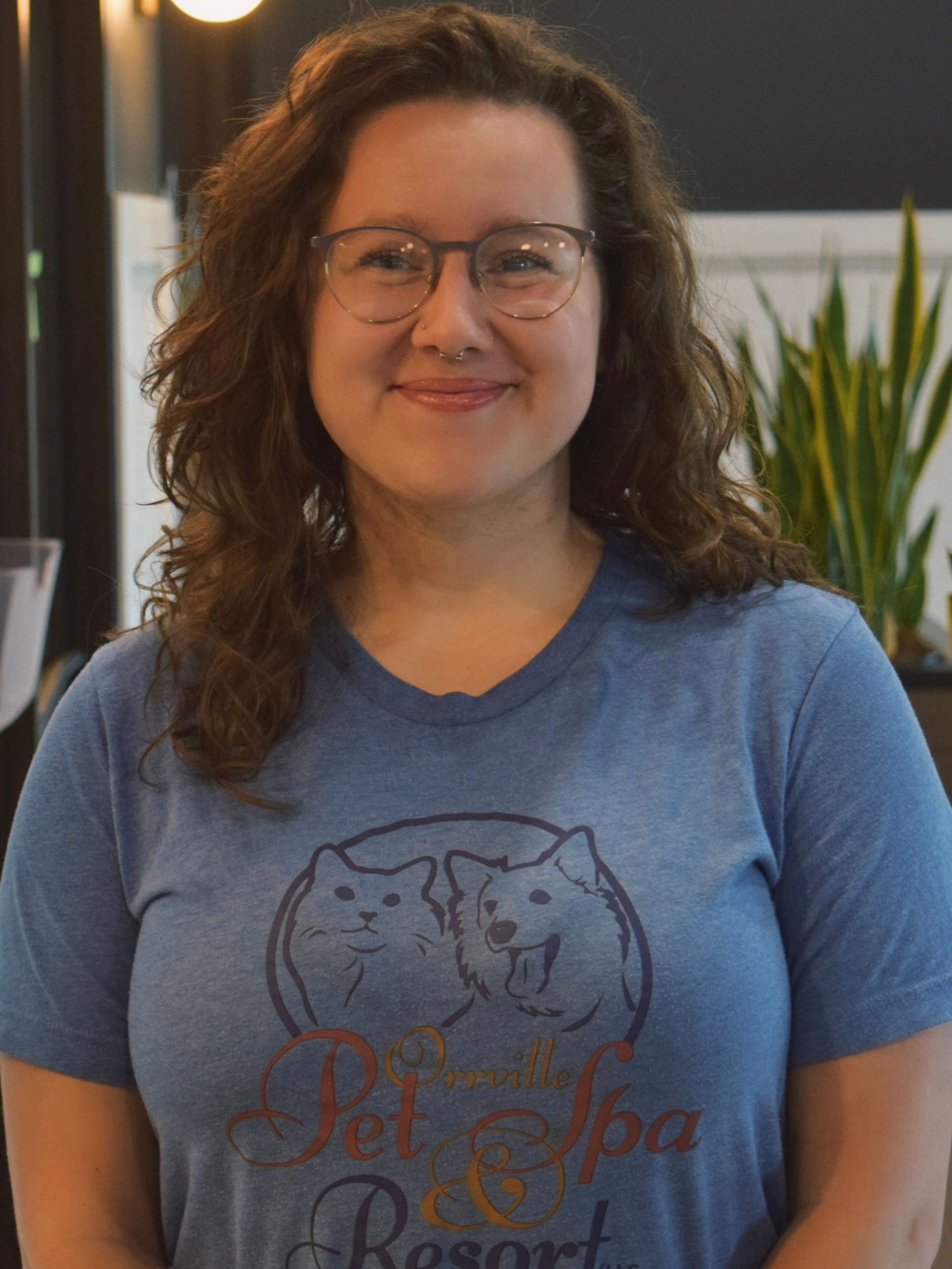 Smiling young woman with curly brown hair and glasses in a blue t-shirt, standing indoors with plants in the background.