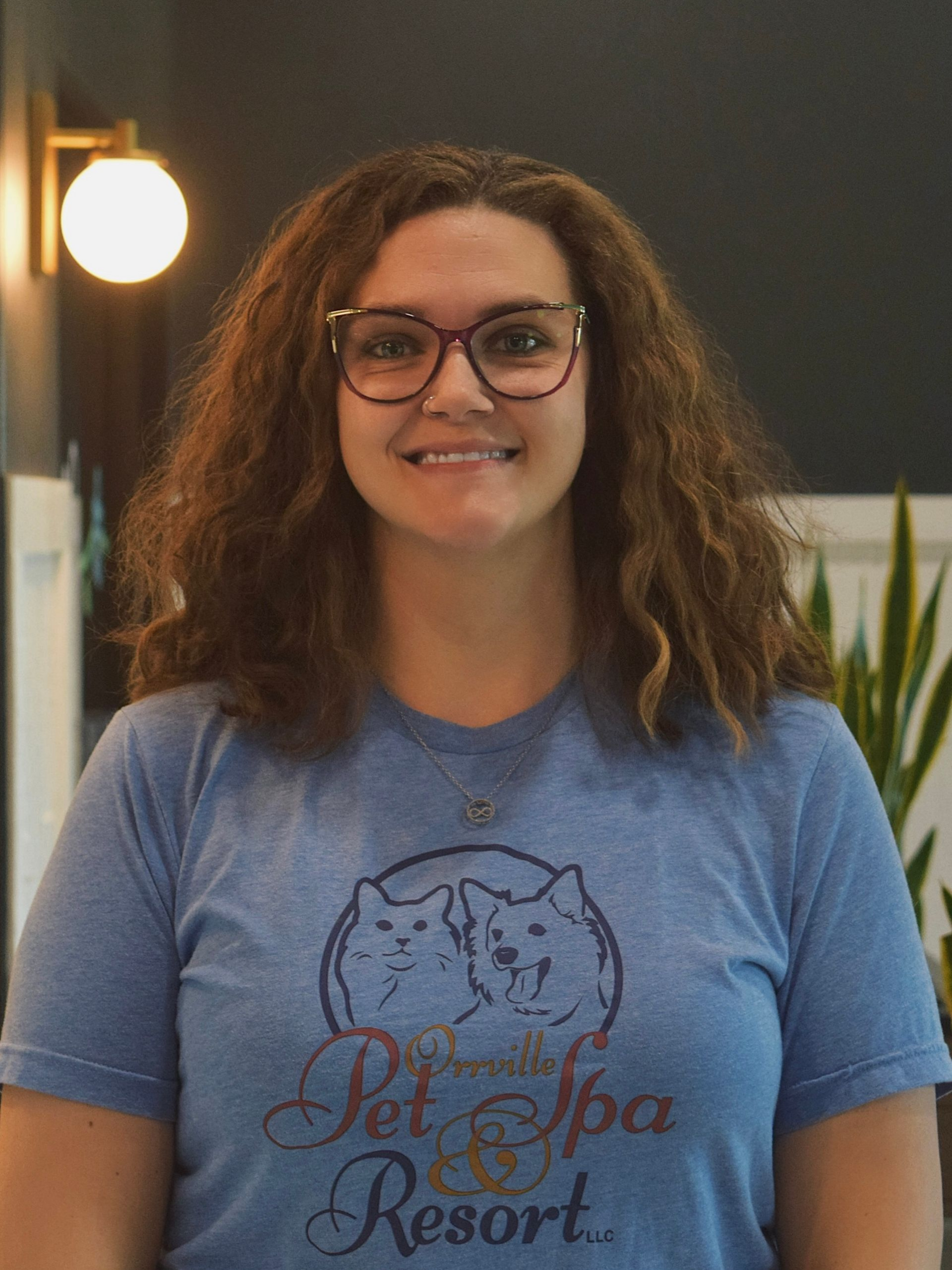 Woman with curly hair, glasses, and blue shirt smiles at the camera in an office setting.