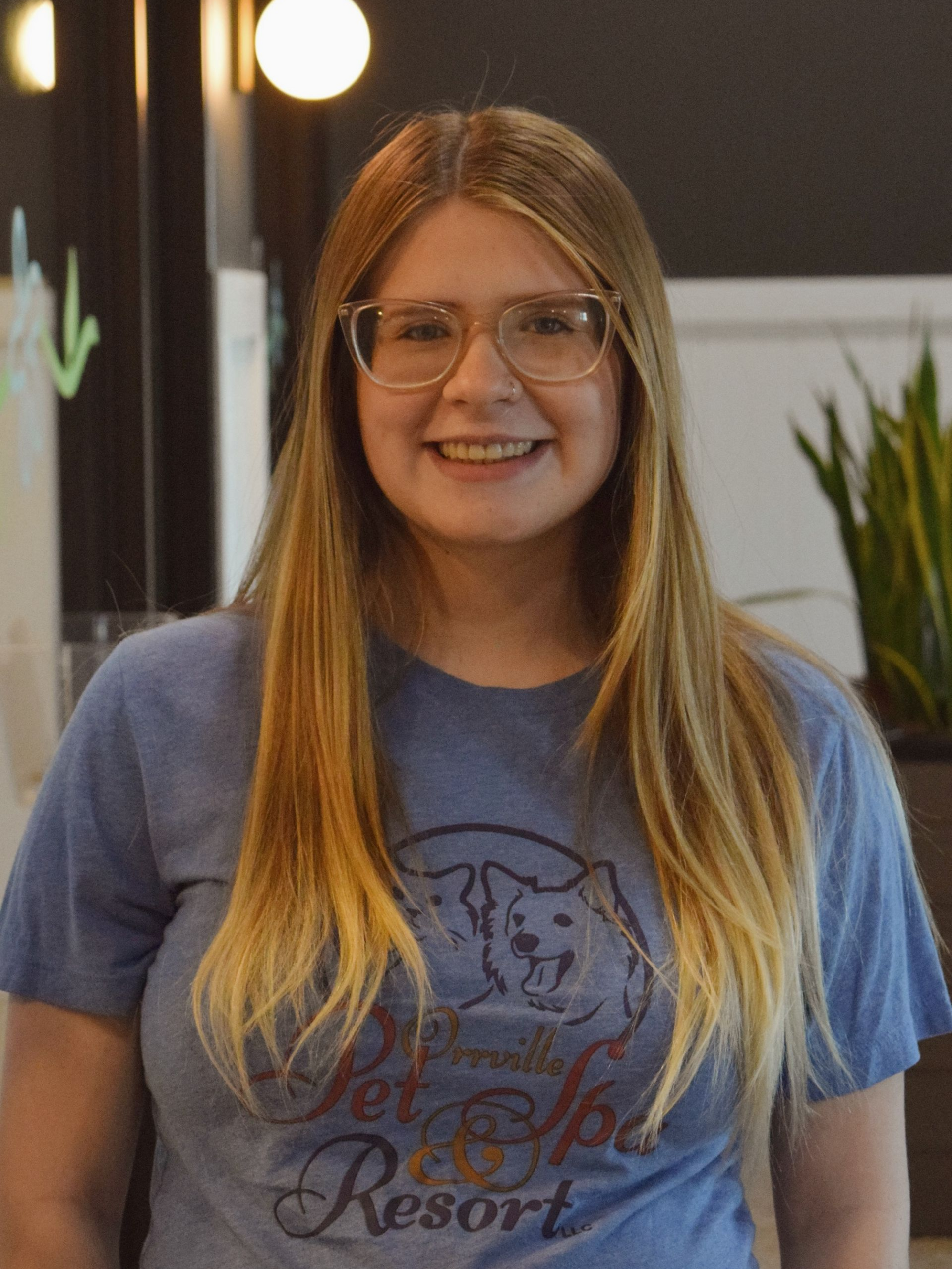 Woman with blonde hair wearing a blue hoodie and glasses sits in a wooden chair, smiling. She is indoors near plants and a black wall.