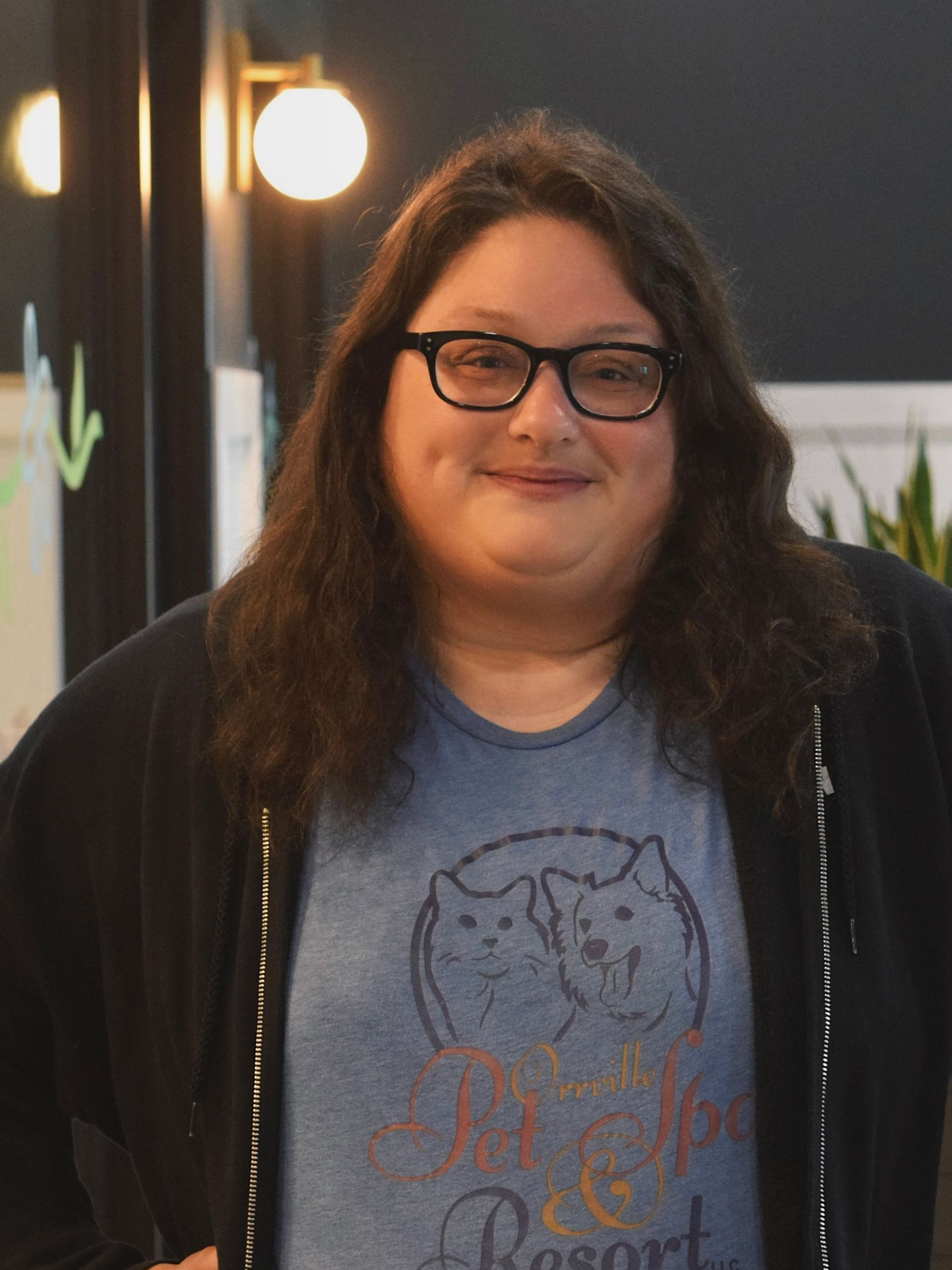 Woman with glasses and a cat shirt smiles indoors. Plants in the background. Pink purse strap across the front.
